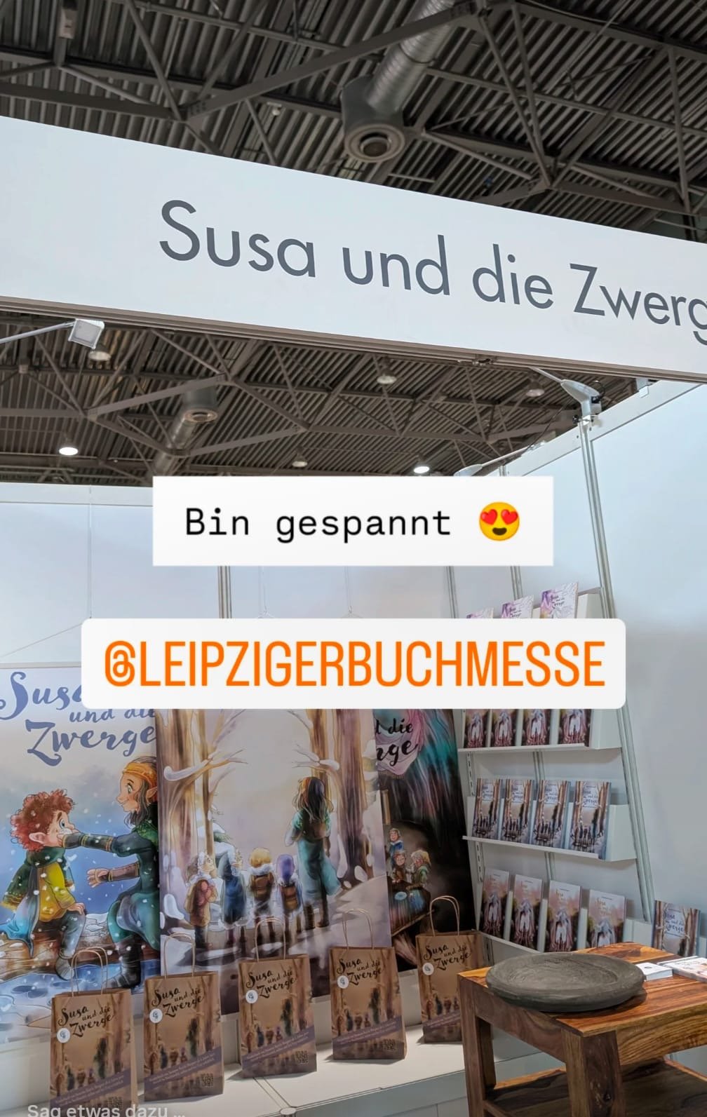 Buchstand auf der Leipziger Buchmesse mit einem großen Banner, das 'Susas und die Zwerge' zeigt, darunter eine Illustration von Zwergen und Charakteren, sowie mehrere Bücher und Poster des Buches. Eine Person macht ein Foto des Stands.