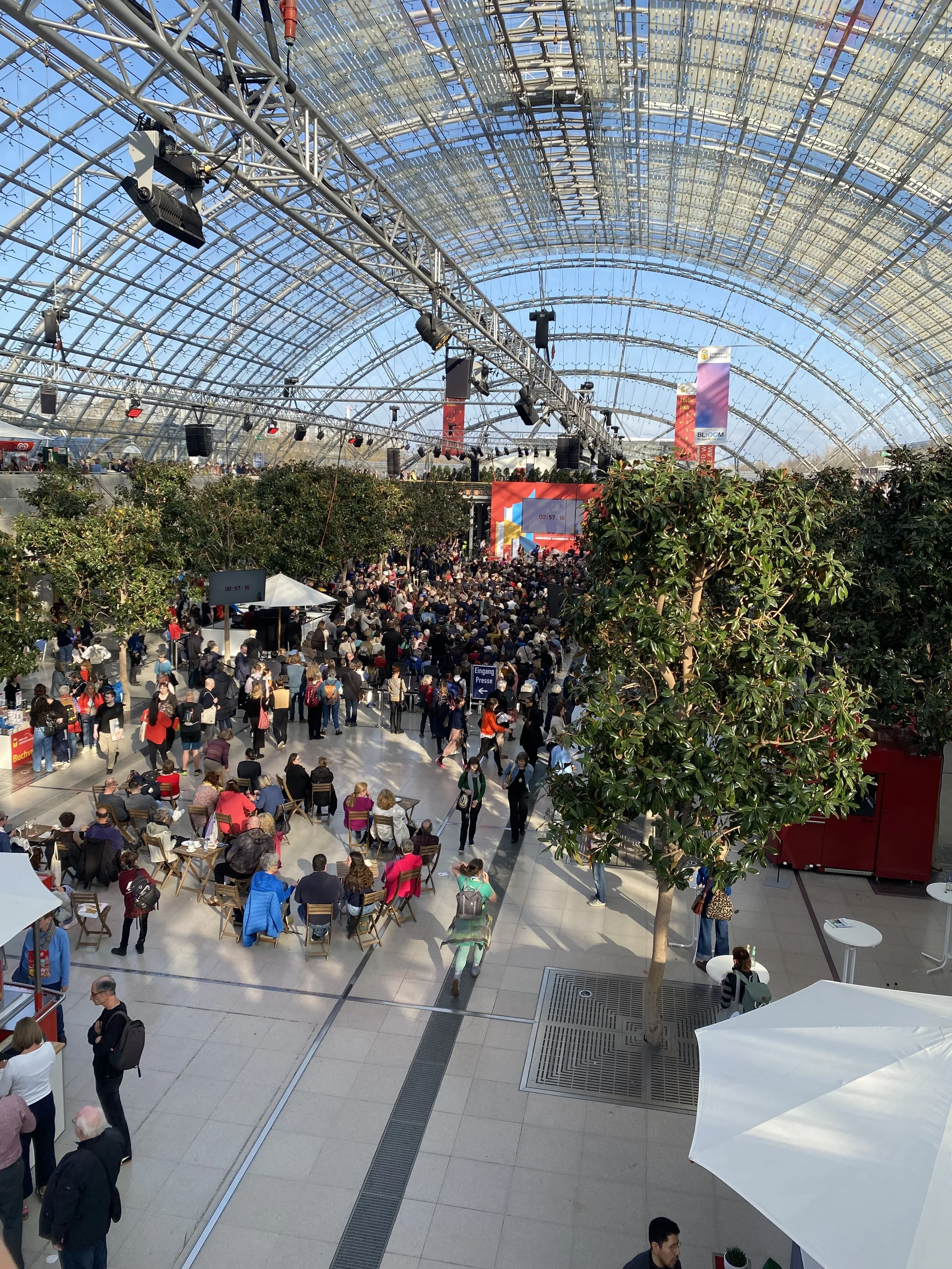 Innenansicht eines großen Veranstaltungssaals mit vielen Zuschauern, einer Bühne im Hintergrund, einer großen LED-Leinwand, Bäumen im Raum, und einer transparenten, gewölbten Glasdecke im Dach des Gebäudes.