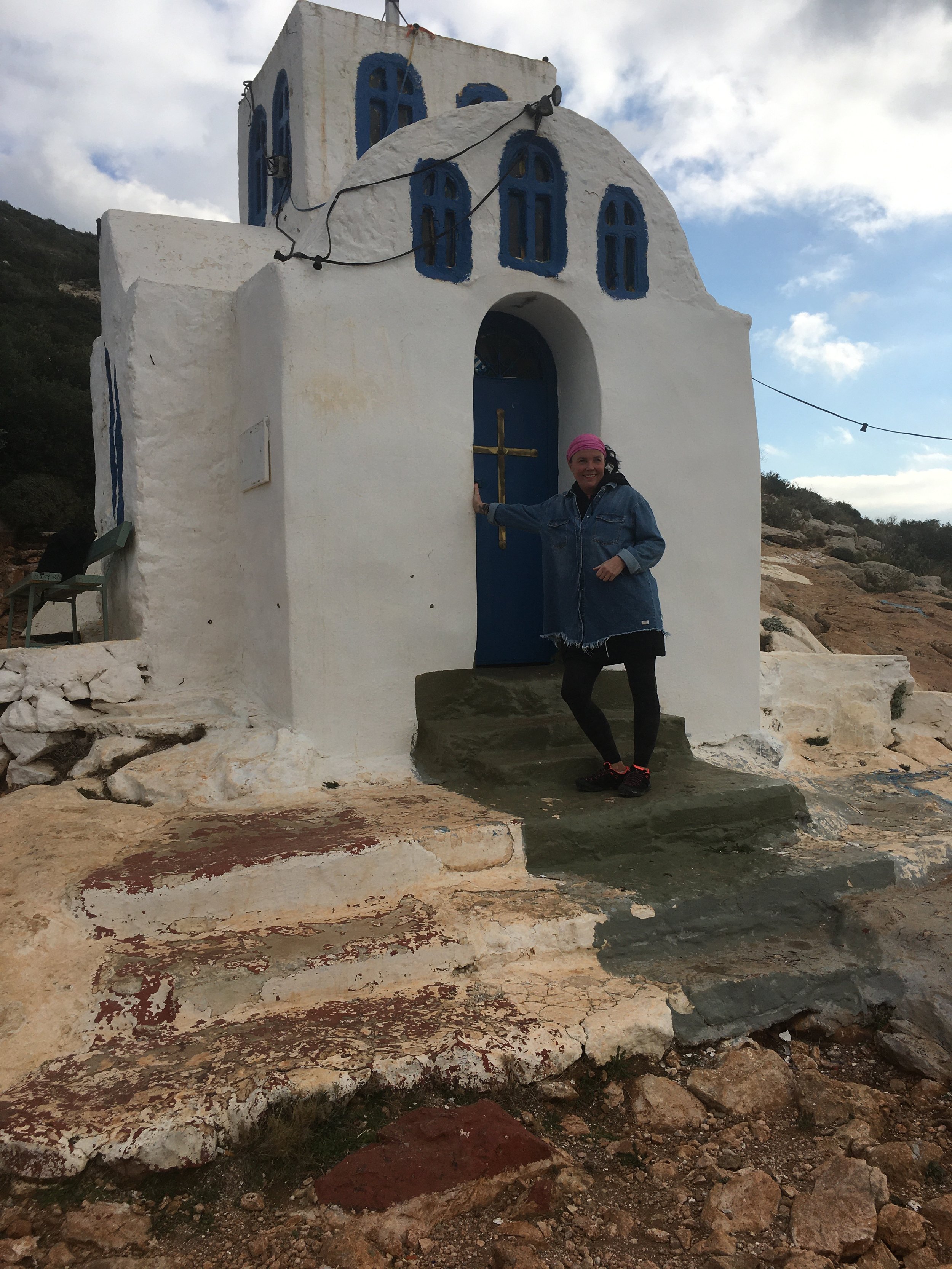 Ein weißes kleines Kirchengebäude mit blauen Fenstern und einer blauen Tür, daran eine Person mit pinker Bandana und Jeansjacke, die die Tür berührt, im Freien, bei bewölktem Himmel auf einer felsigen Erde.