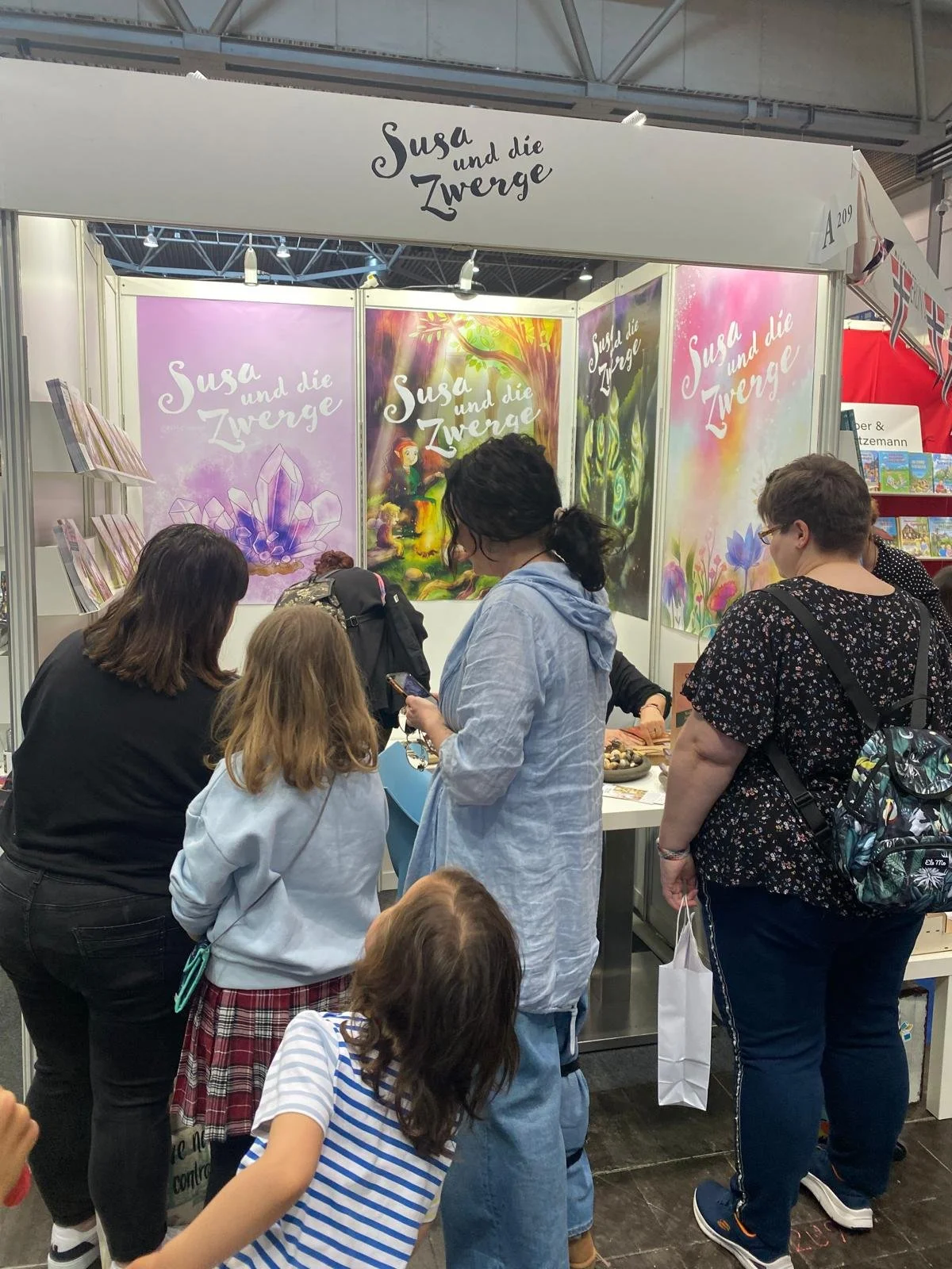Stoffstand mit Büchern und bunten Postern beim Messestand "Susa und die Zwerge", Besucher stehen und schauen.