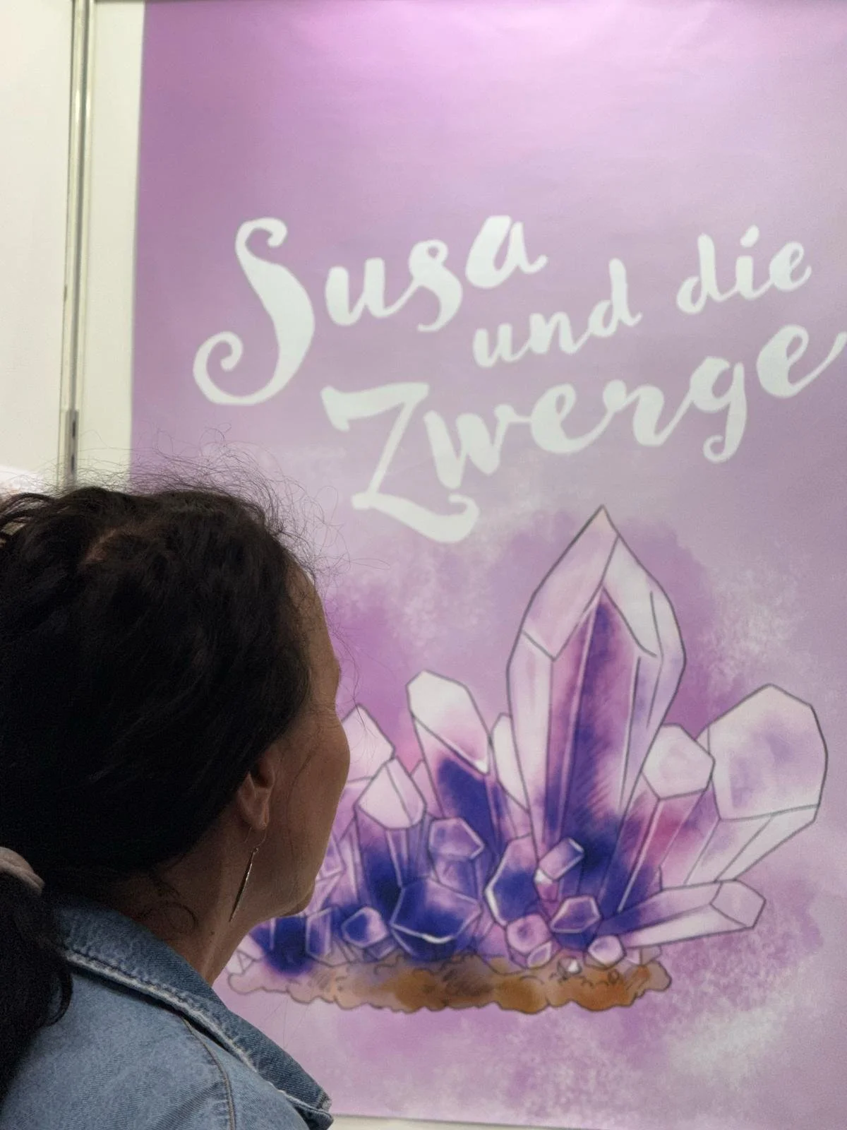 Das Bild zeigt eine Person mit dunklen Haaren, die vor einem Poster steht. Das Poster hat einen lila Hintergrund und zeigt einen großen Kristall mit mehreren kleineren Kristallen rundherum. Oben im Poster steht in weißen, verspielten Buchstaben: "Susza und die Zwerge".