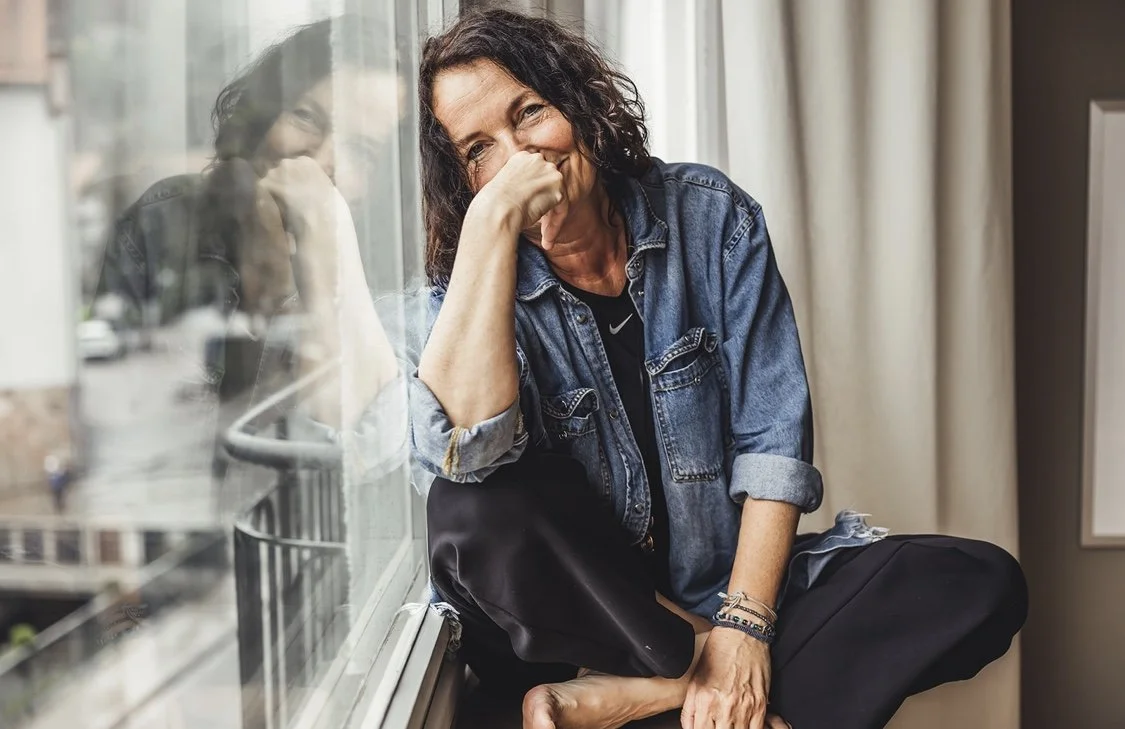 Frau mit dunklen, lockigen Haaren, die lachend neben einem Fenster sitzt, die Hand vor das Gesicht hält, im Hintergrund eine Stadt.