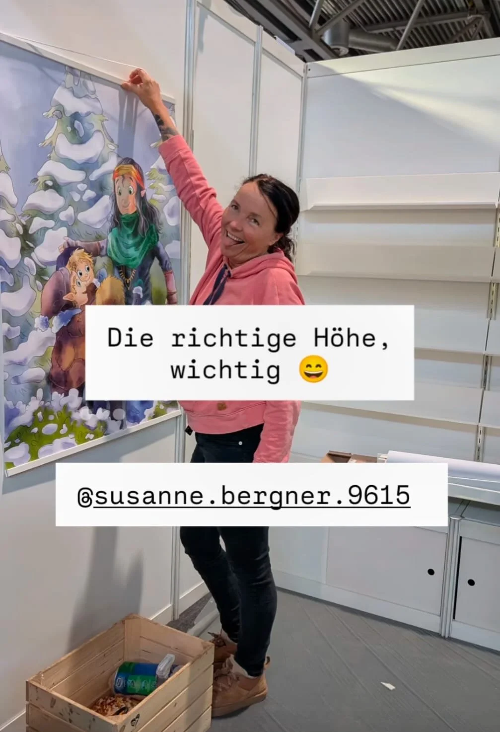 Eine Frau hängt ein buntes Kinderposter mit einer Märchenfigur an einer Messwand. Sie lächelt und scheint stolz auf die Platzierung. Es gibt einen Holzkasten mit Getränken und Snacks auf dem Boden davor.