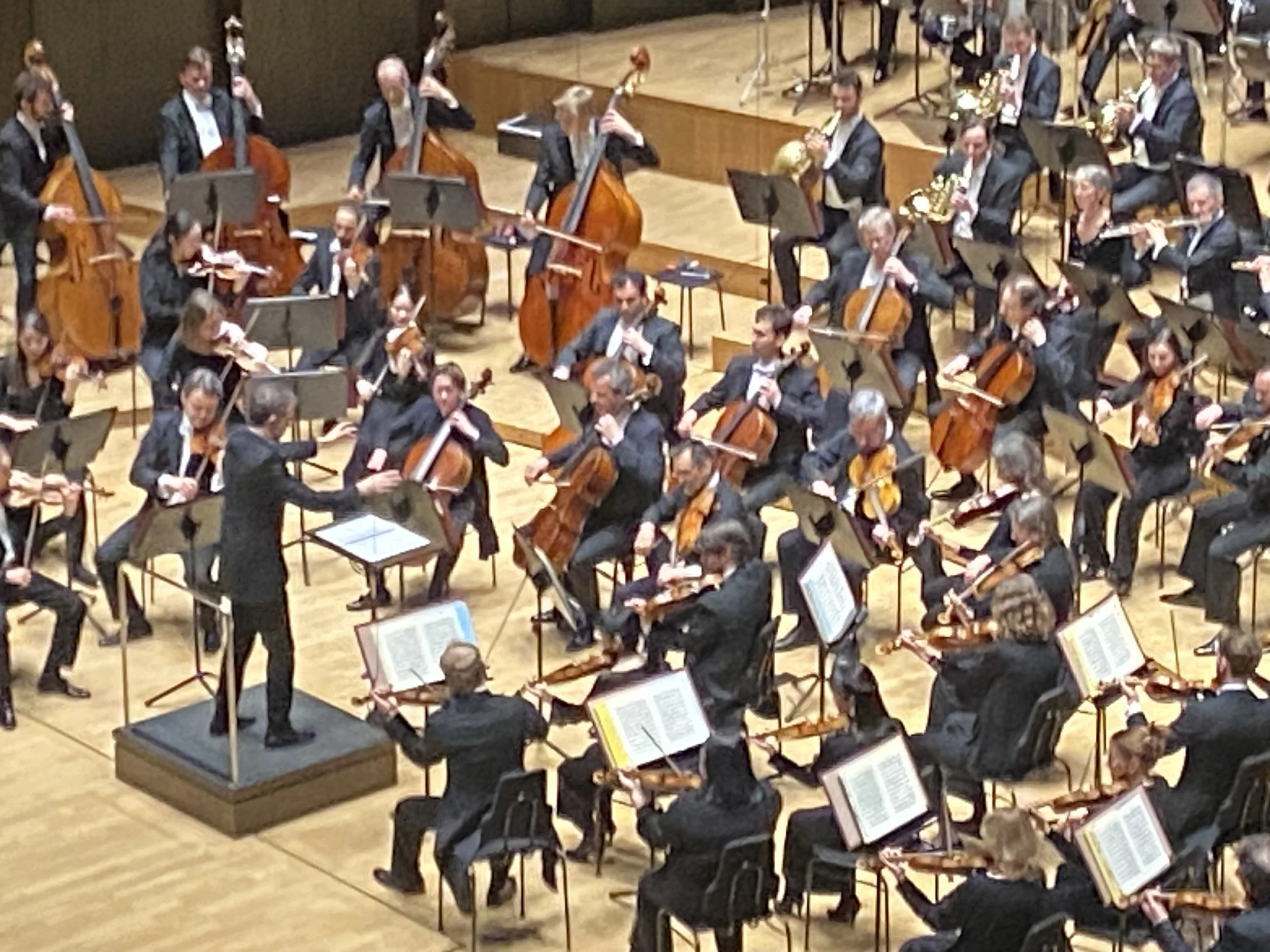 Ein Orchester mit Musikern auf einer Bühne, die verschiedene Instrumente spielen, darunter Geigen, Celli, Trompeten und Flöten. Der Dirigent steht vorne auf einem Pult, der Rufenseinheit und der Aufbau deuten auf eine klassische Aufführung hin.
