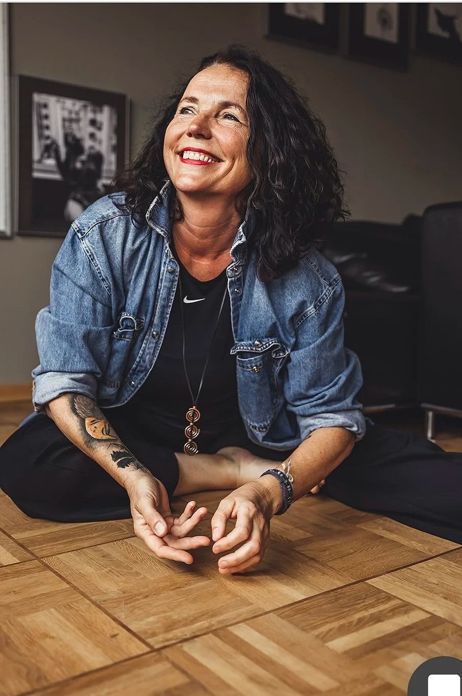 Eine lachende Frau mit schwarzen, lockigen Haaren sitzt auf dem Boden, trägt eine Jeansjacke und eine schwarze T-Shirt mit Nike-Logo, zeigt ein Tattoo am linken Arm, trägt Schmuck und sitzt in einem Raum mit Holzdielenboden und Wandbildern im Hintergrund.