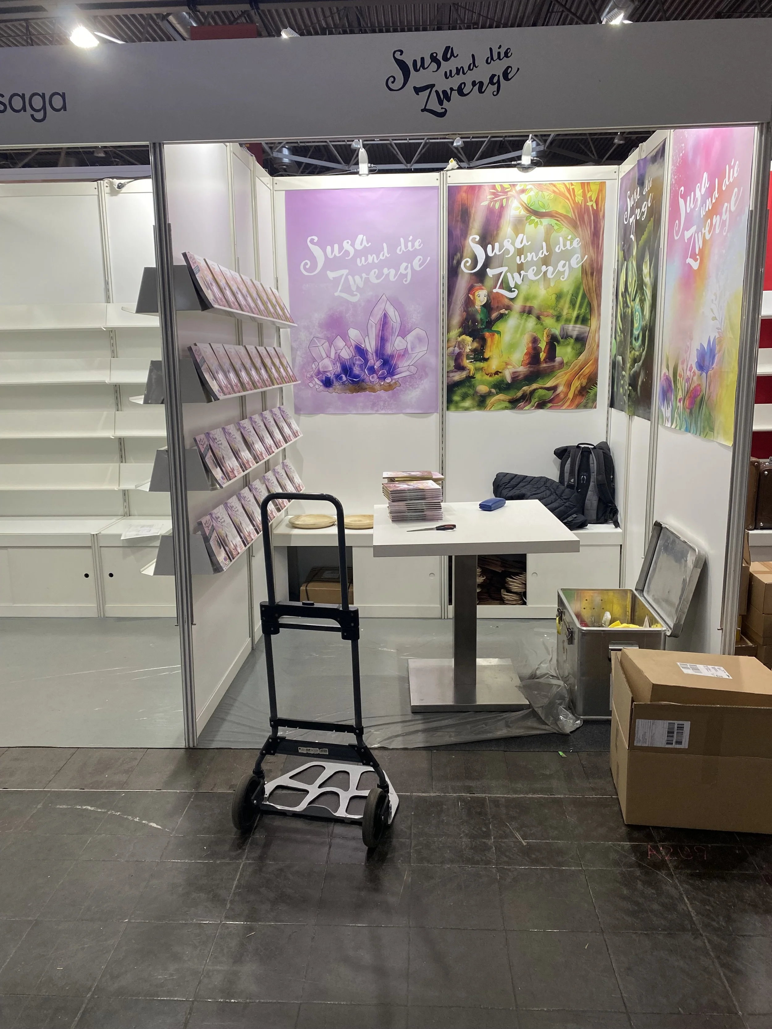 Messestand mit Bannern der Kinderbuchreihe 'Susa und die Zwerge', eine Tafel mit Broschüren, ein Packstück, ein Kinderwagen, und Faltkartons auf Boden.