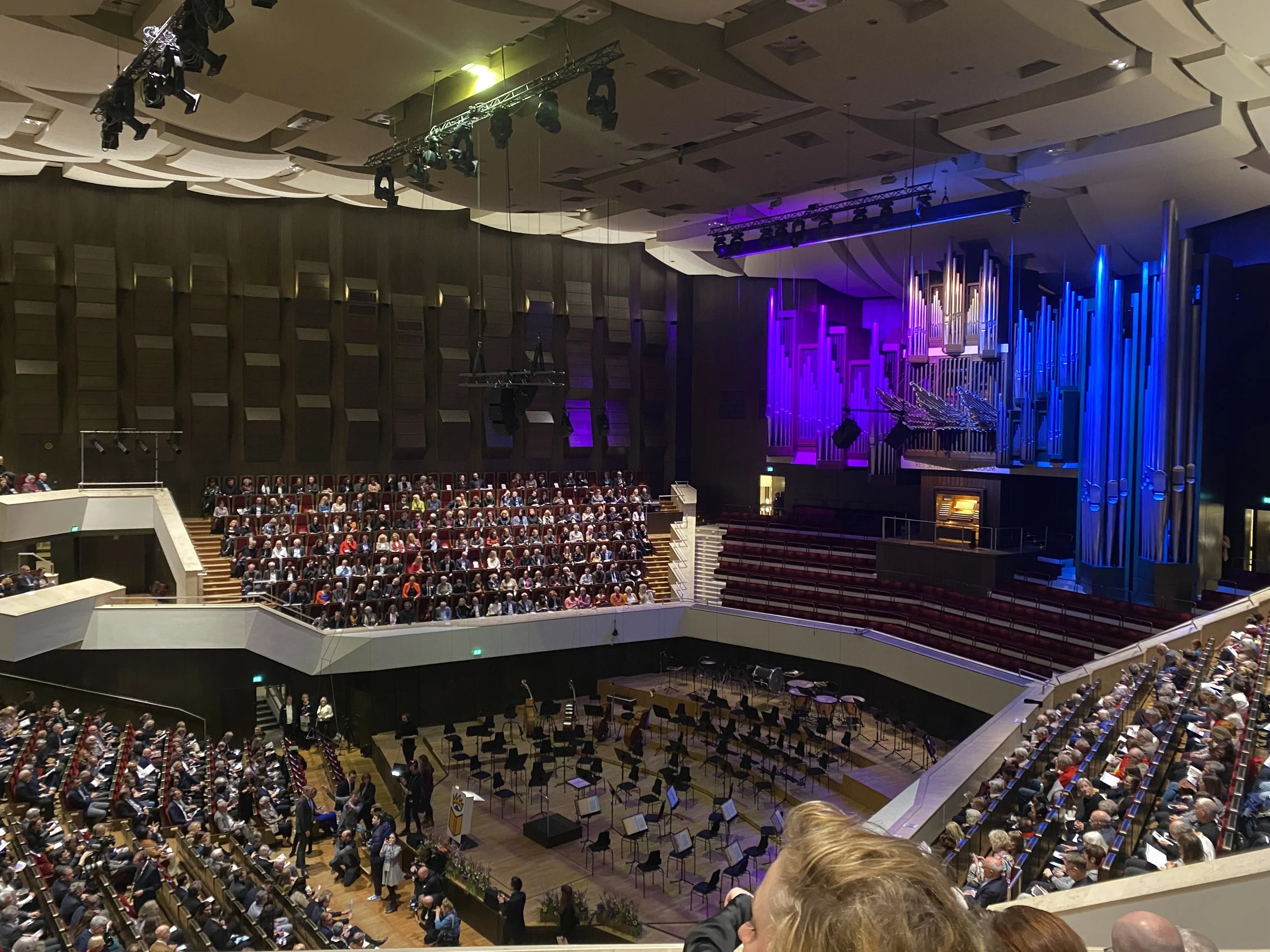 Ein großes Konzertsaal mit einem Orchester auf der Bühne und vielen Zuschauern im Publikum, lila und blaue Beleuchtung, moderne Architektur mit Holzverkleidung und balgigen Türen.