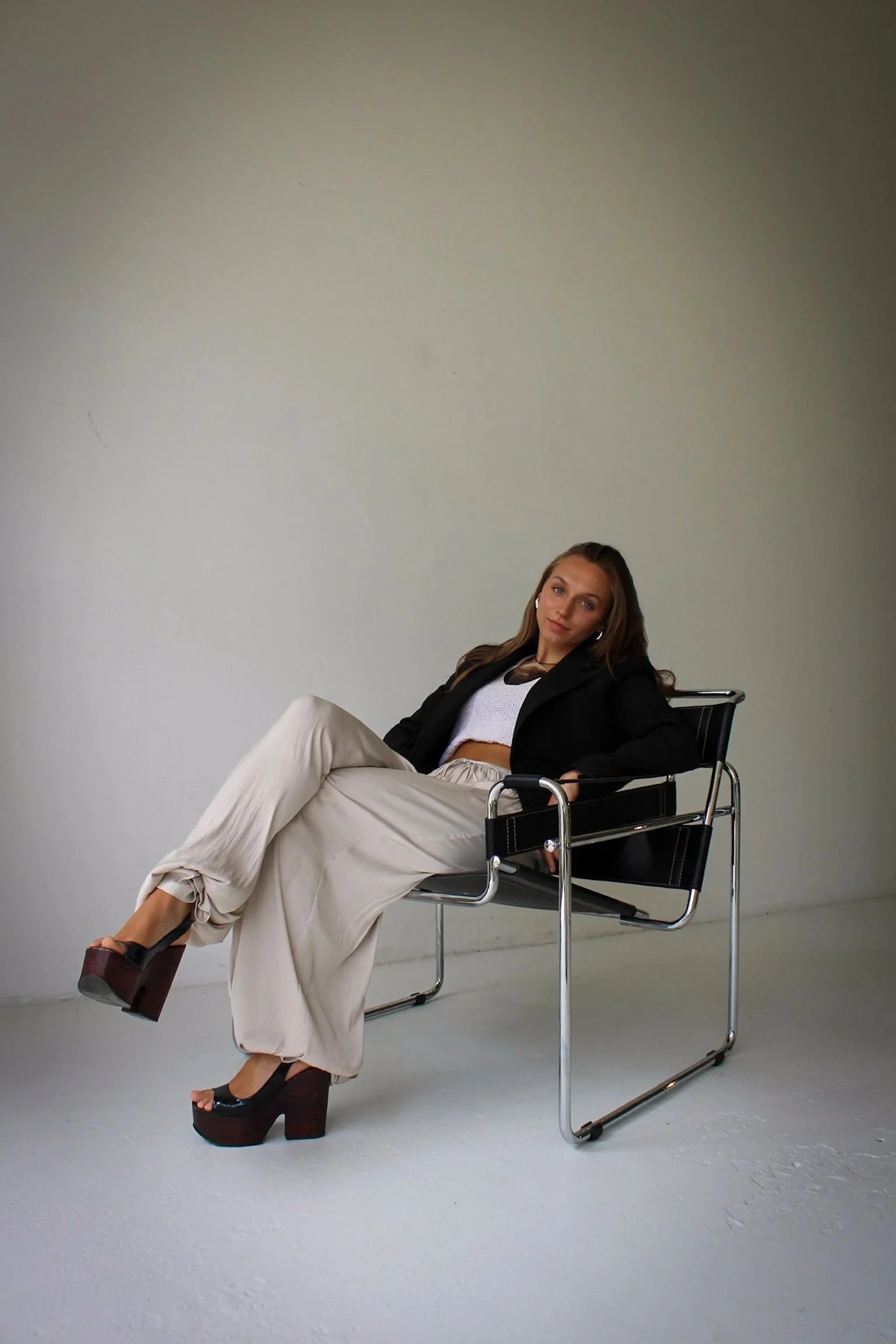 A young woman with long hair sitting in a modern black chair against a plain white wall, wearing a black blazer, white crochet top, white loose pants, and high platform sandals.