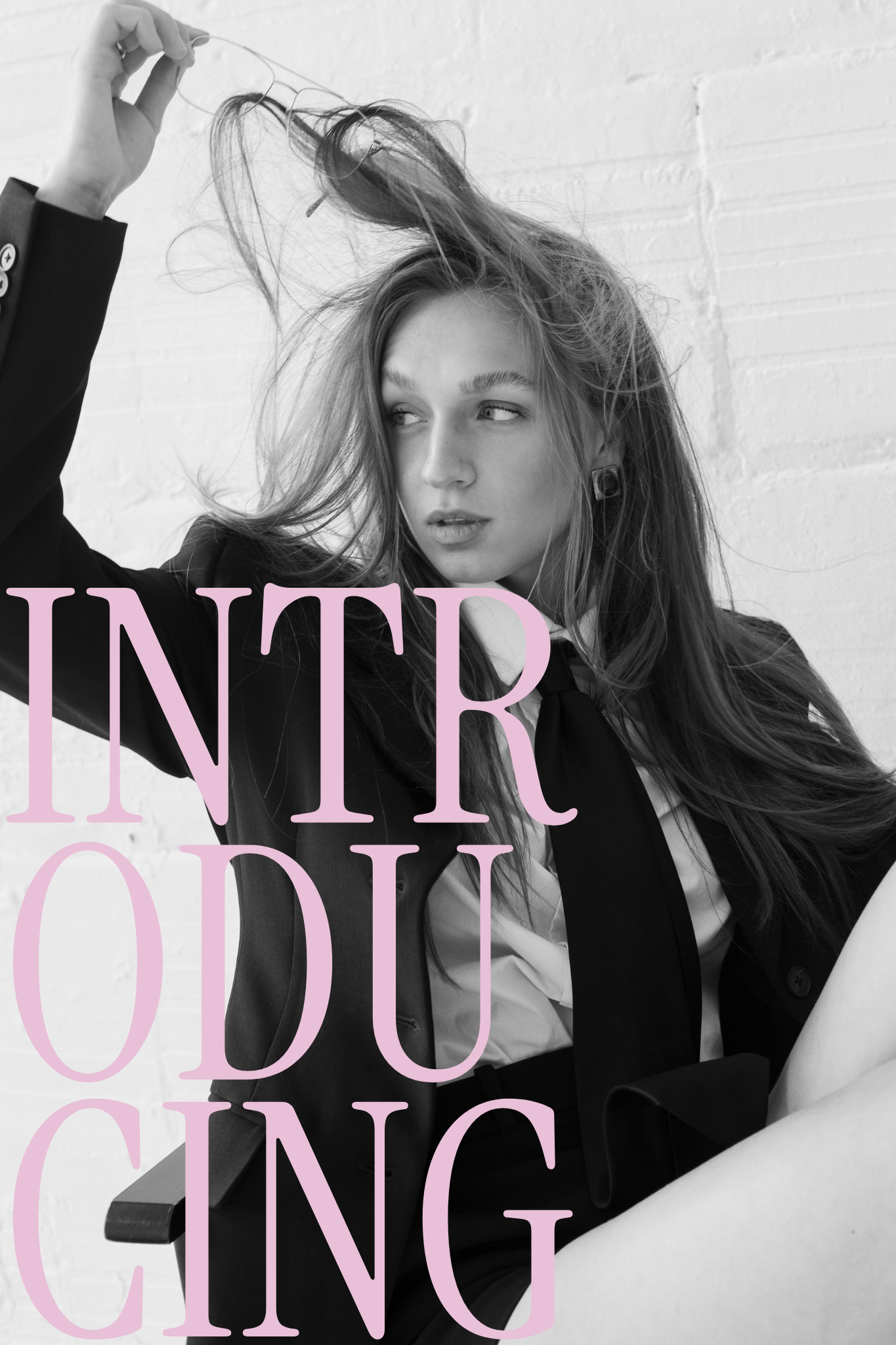 A young woman with long hair is sitting against a white brick wall, looking to the side while holding her hair with one hand. She is wearing a black blazer over a light shirt. The image is in black and white with large, light purple text overlaying part of the image.