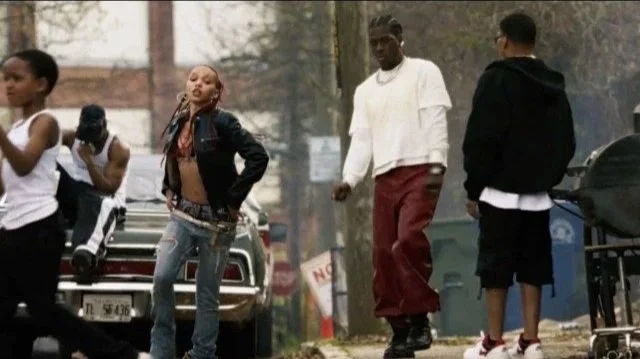 Group of young people walking in an urban neighborhood, some wearing casual clothing, with cars and trees in the background.