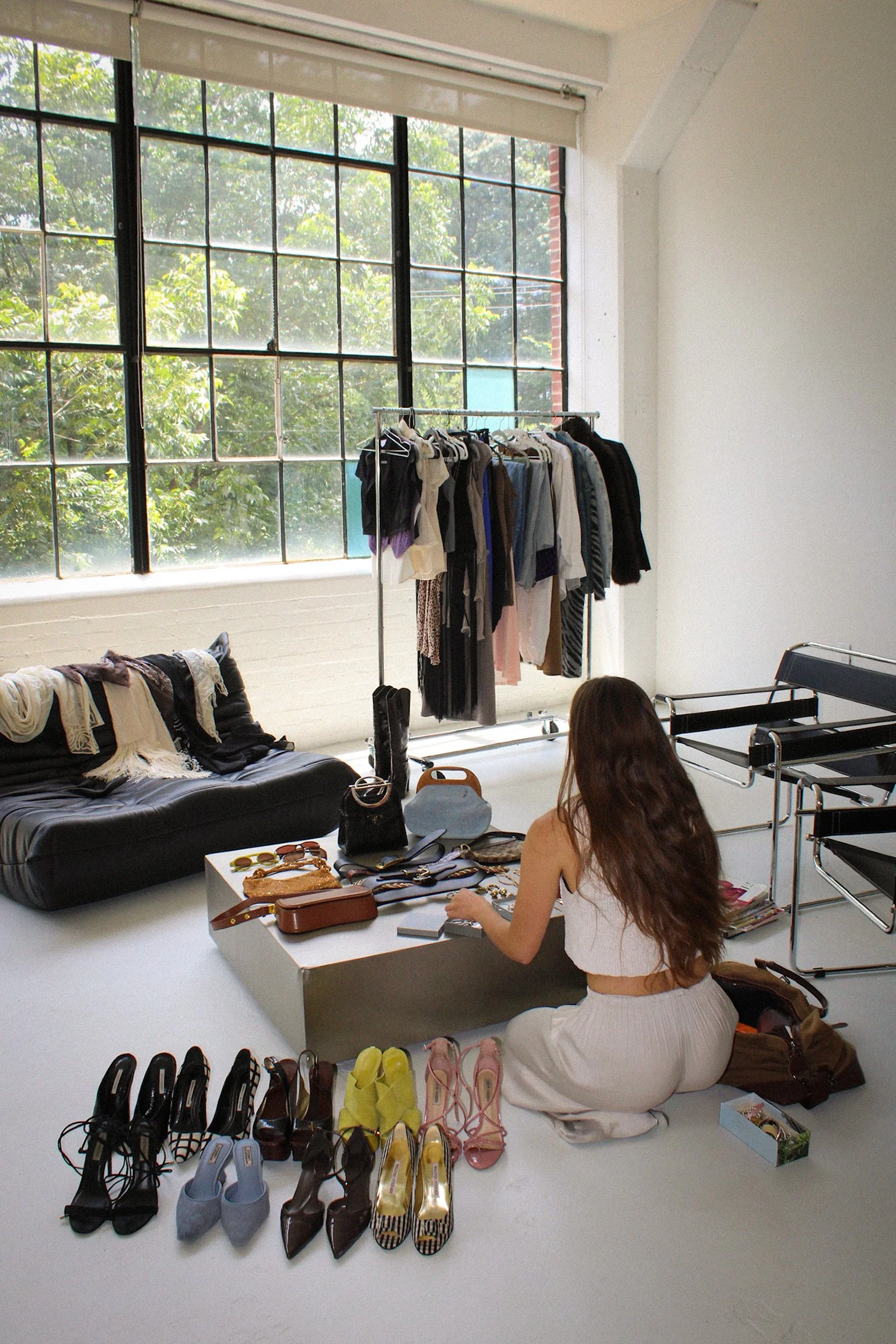 A woman with long brown hair wearing a white top and white skirt, sitting on the floor in a bright room with large windows. She is surrounded by shoes, accessories, and jewelry on a low white table and the floor. There is a black leather couch with clothes on it, and a clothing rack with hanging clothes in the background. The room appears to be a studio or dressing area.