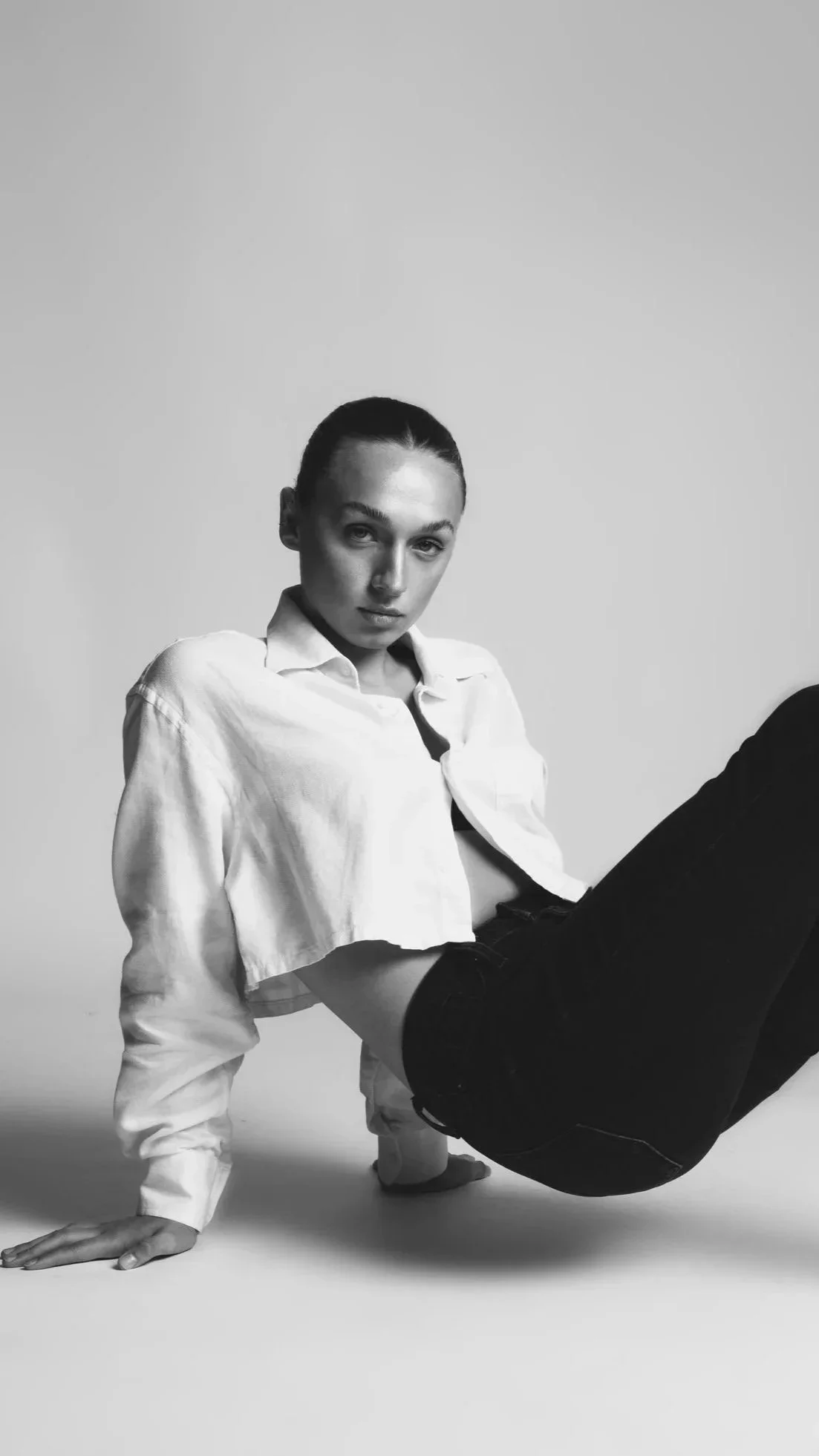 Black and white photo of a woman in a white shirt and black pants, posed on the floor in a dance or yoga pose, looking at the camera.