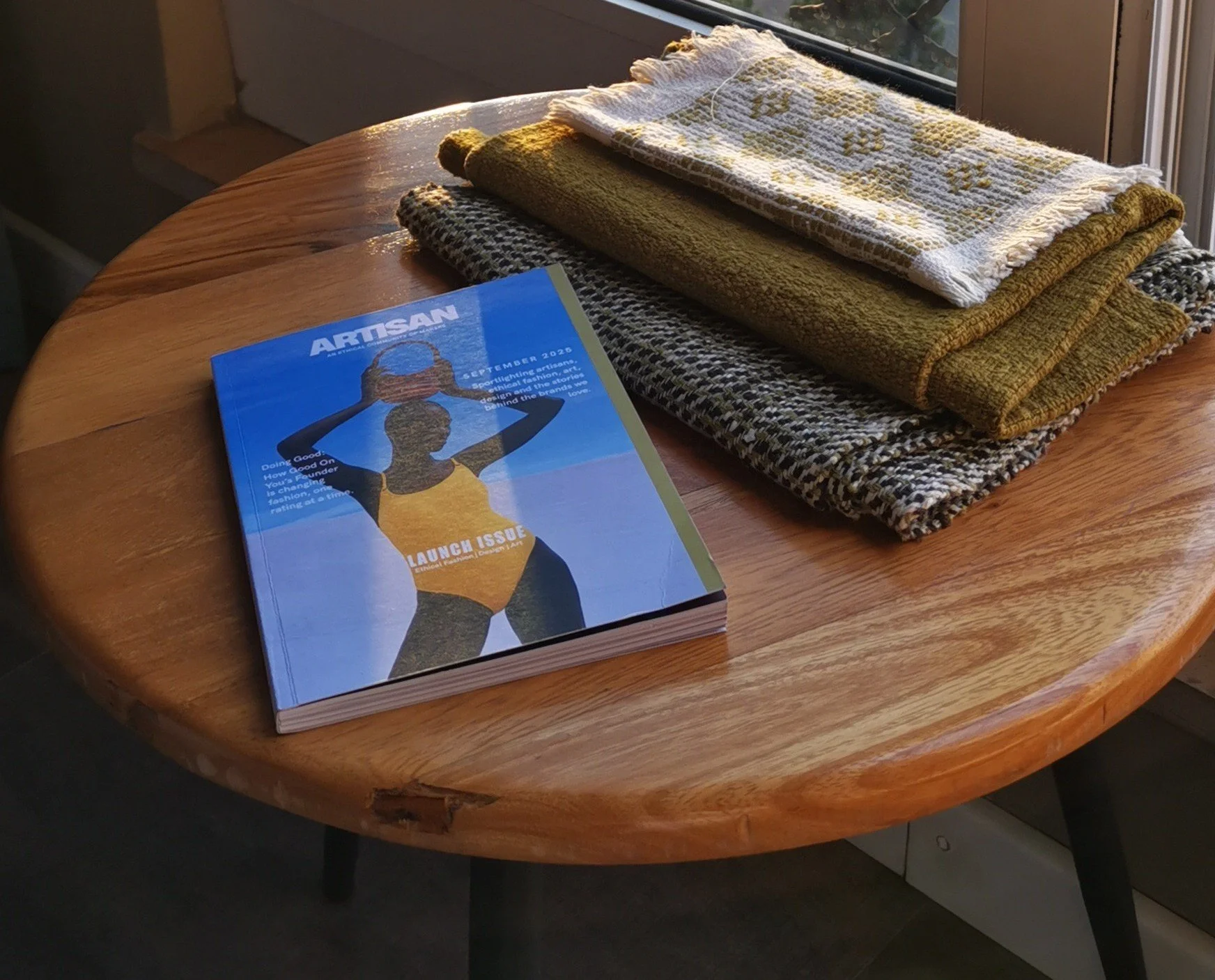 Wooden table with a magazine titled "ARTISAN" and a group of folded towels in various colors and patterns, positioned near a window with sunlight.
