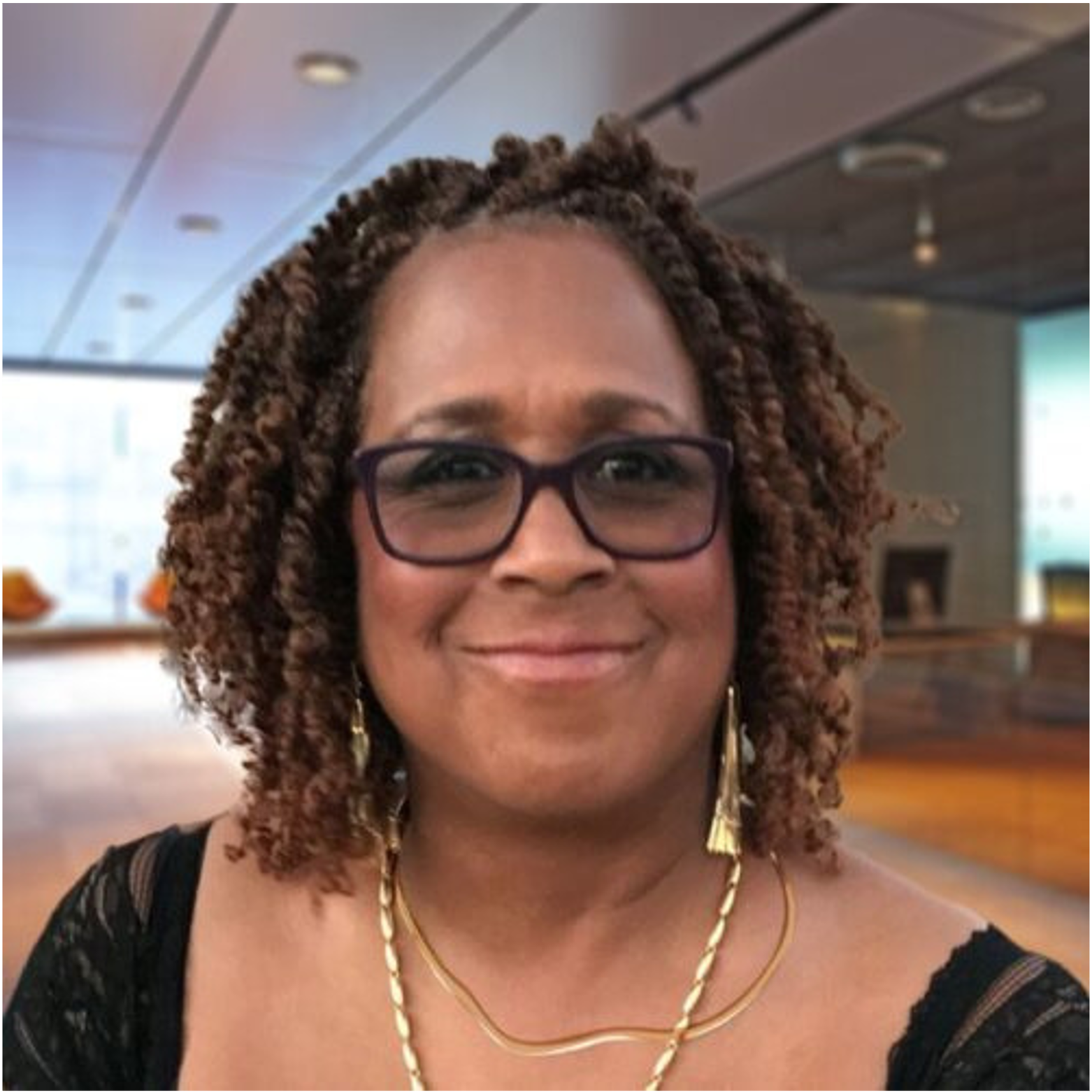 A woman with curly brown hair, wearing glasses, gold earrings, and jewelry, smiling in an indoor setting.