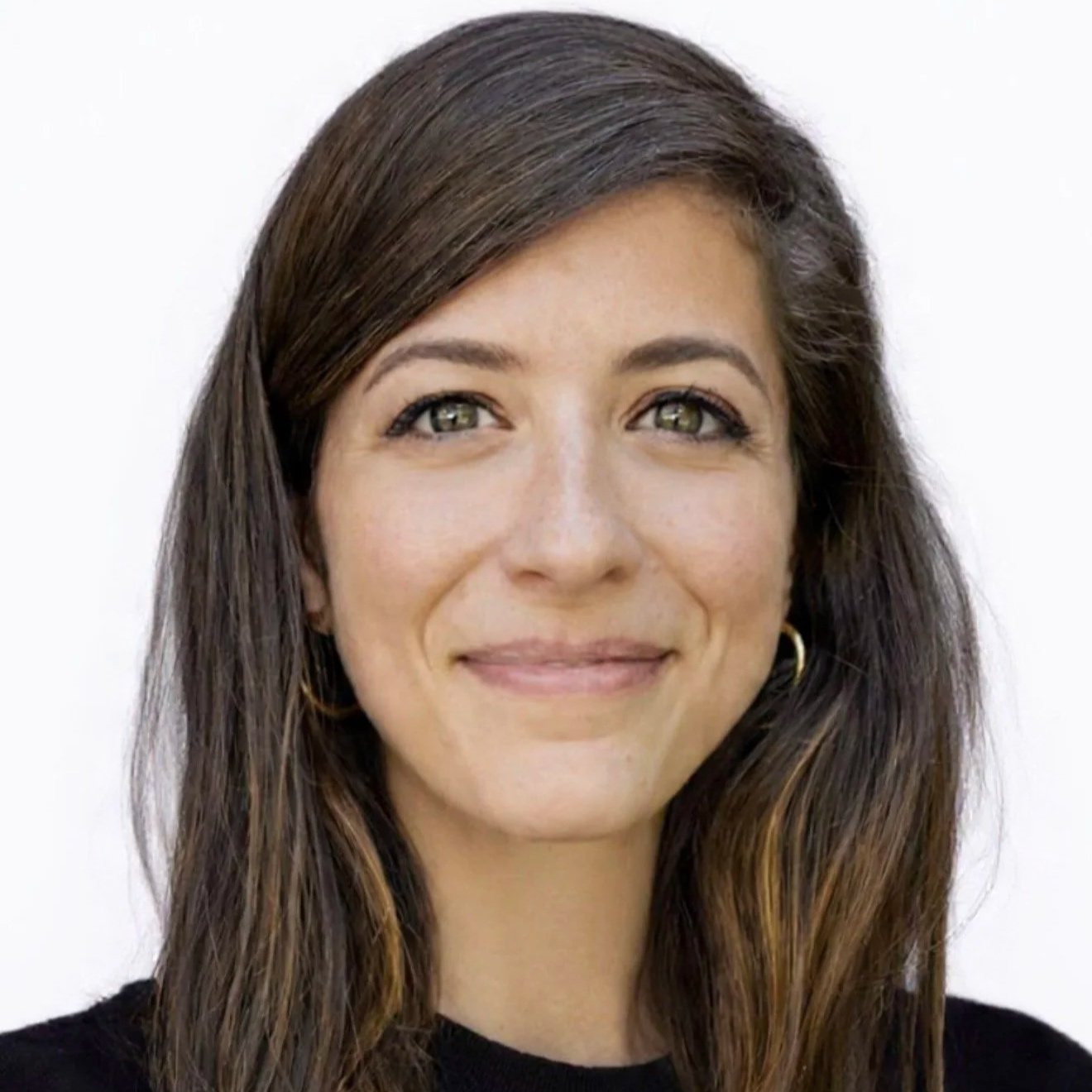 Headshot of a woman with long wavy brown hair and hazel eyes, smiling, wearing gold hoop earrings and a black top, against a plain white background.