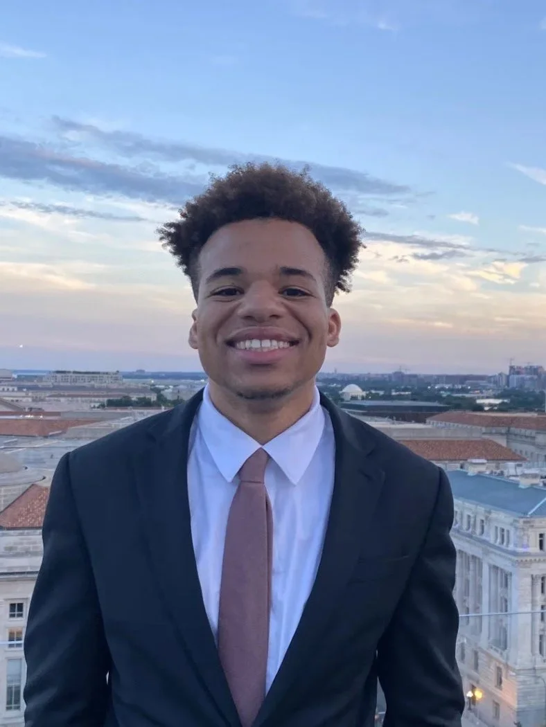 Smiling young man in a black suit and light pink tie standing outdoors on a rooftop, with city buildings and a partly cloudy sky in the background.