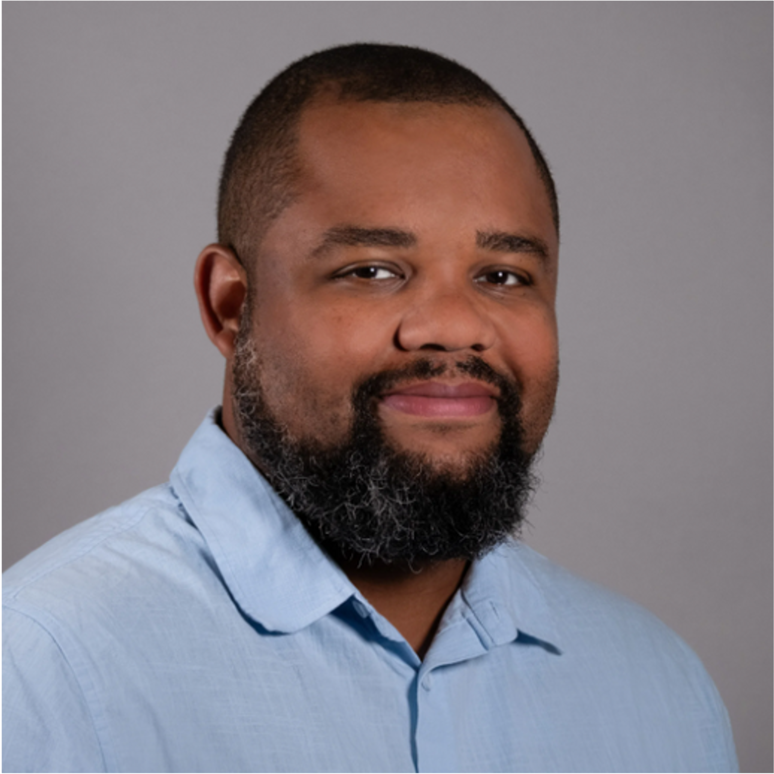 Portrait of a man with a beard and mustache wearing a light blue collared shirt, smiling slightly against a gray background.