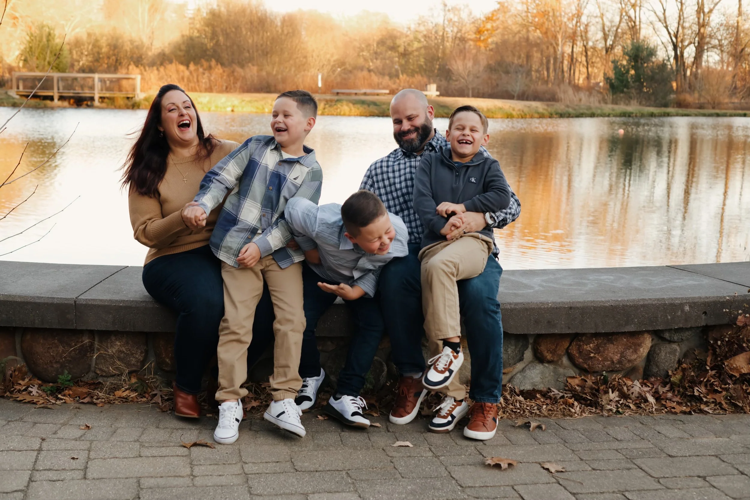 Family of six. 3 children and an adult woman and man laughing and playing by a lake in autumn with trees in the background.