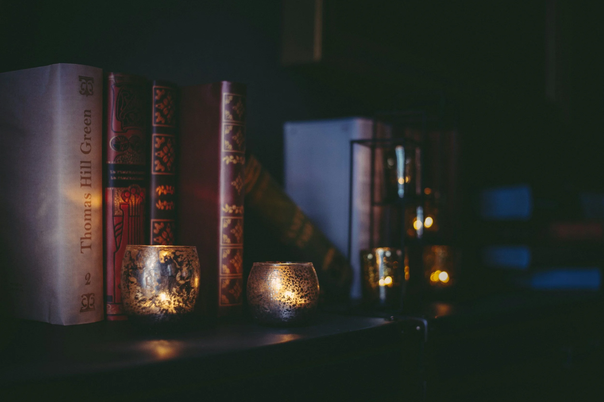 Books on a shelf with decorative candle holders emitting a warm glow, in a dimly lit room with faint reflections.