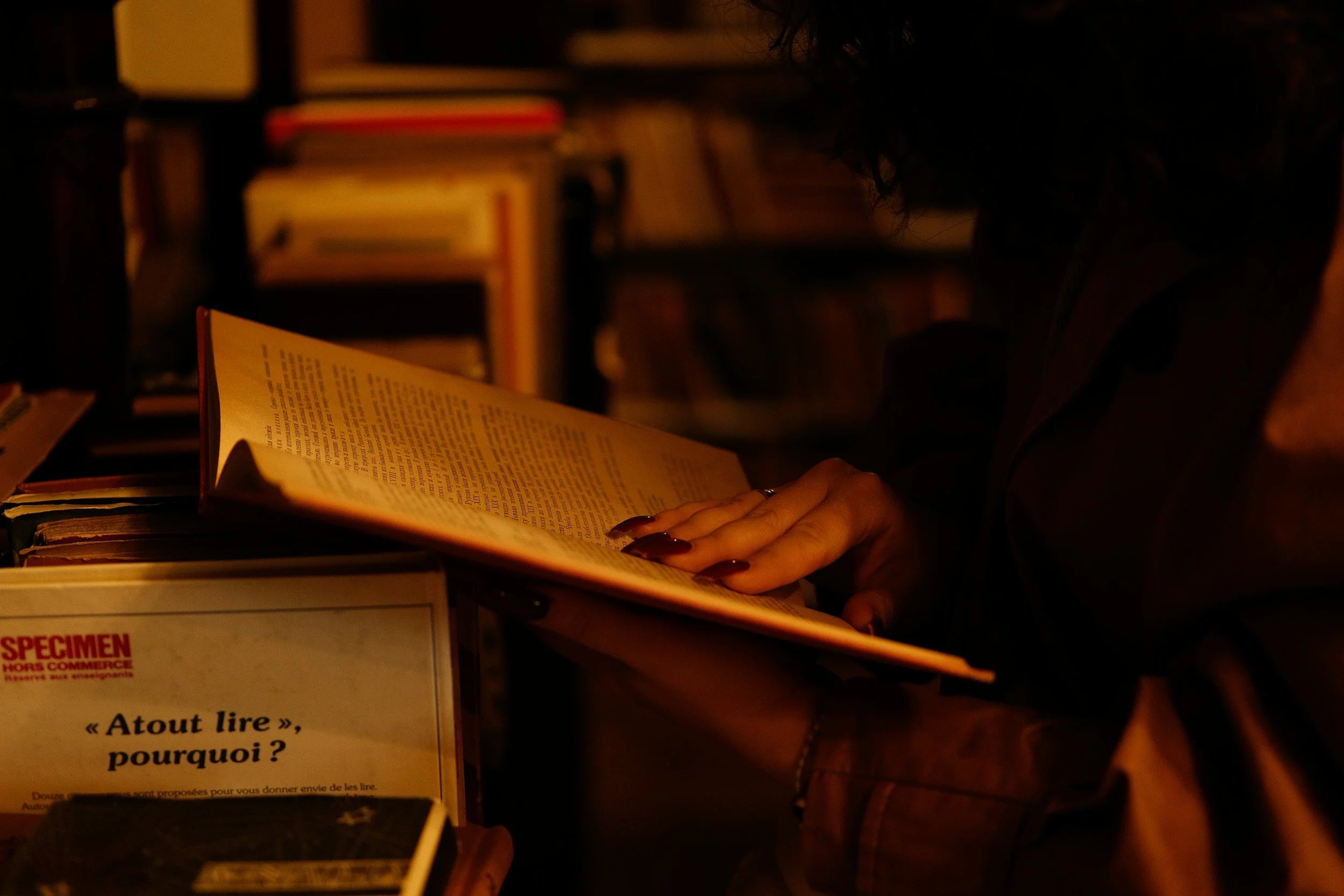 A person with dark, curly hair and dark nail polish reading a book in a dimly lit environment, with shelves of books in the background.