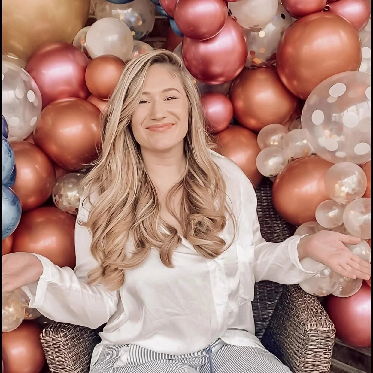 A smiling woman with long blonde hair sitting in front of a large colorful balloon backdrop with metallic and transparent balloons.