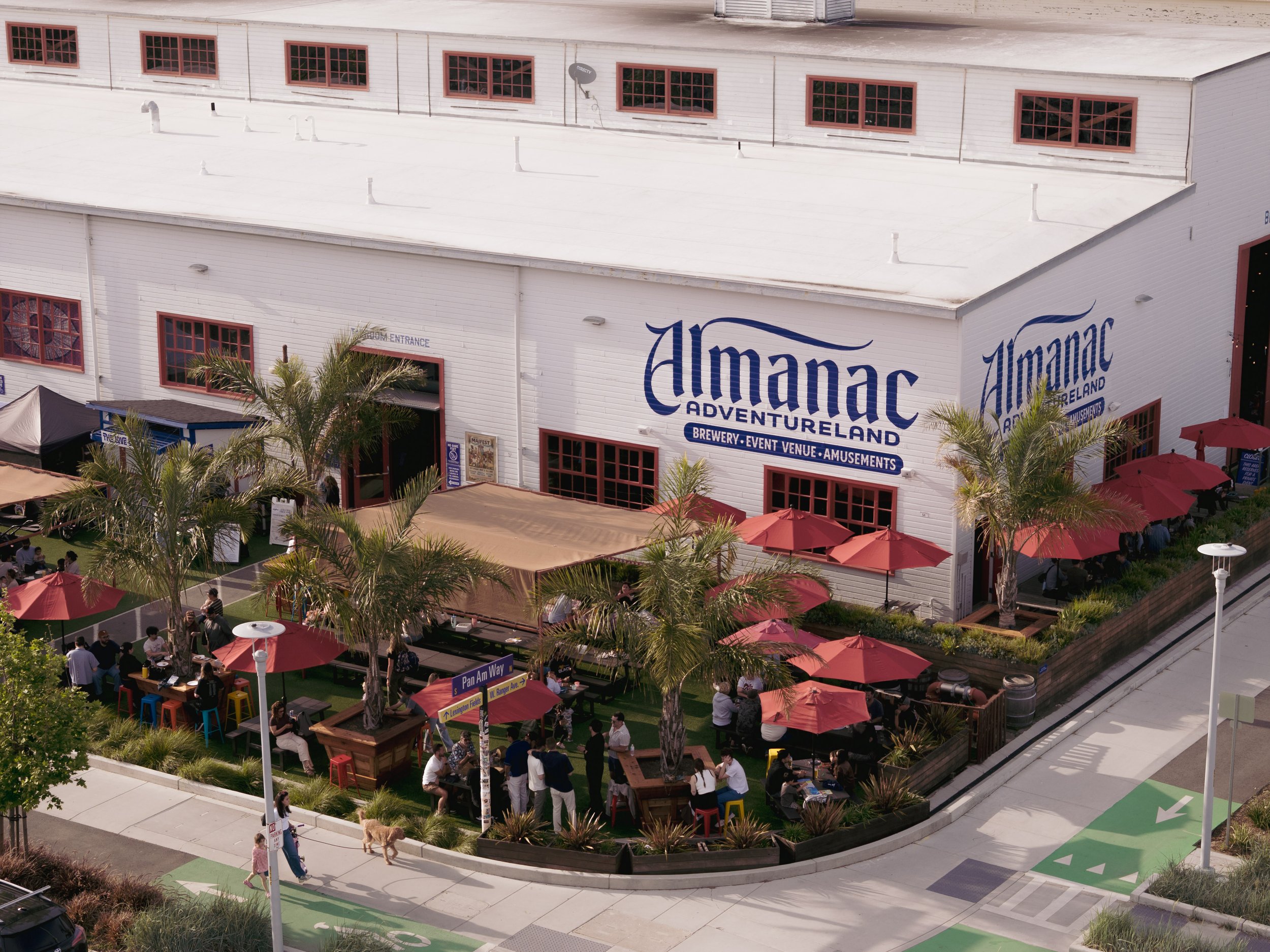 A large white building with red window frames and the words 'Almanac Adventureland' on the side, featuring outdoor seating with red umbrellas and people gathered around, some standing and some sitting, in an outdoor area with palm trees.