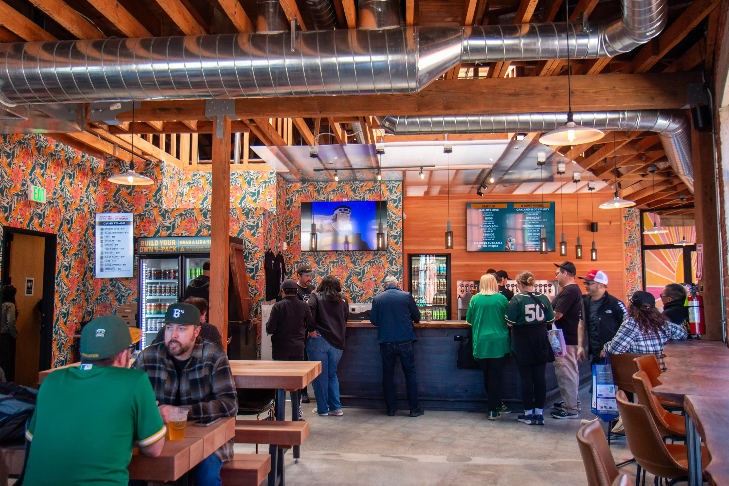 Interior of a busy brewery restaurant with wooden beams, exposed ductwork, floral wallpaper, multiple digital menu screens, and patrons seated at tables.