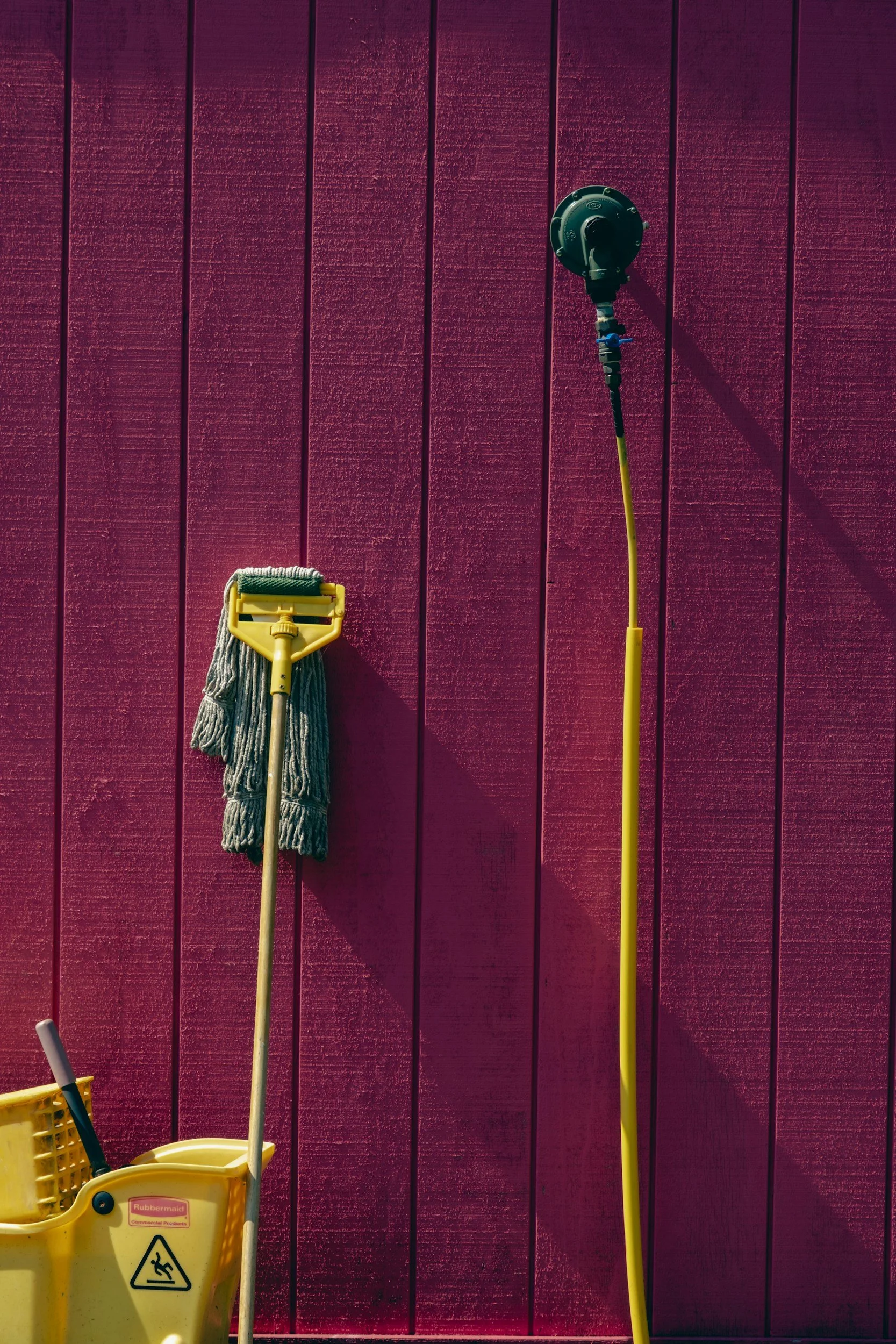Cleaning tools, including a mop, a yellow bucket, and a water spray device, are hanging on a red painted wooden wall.