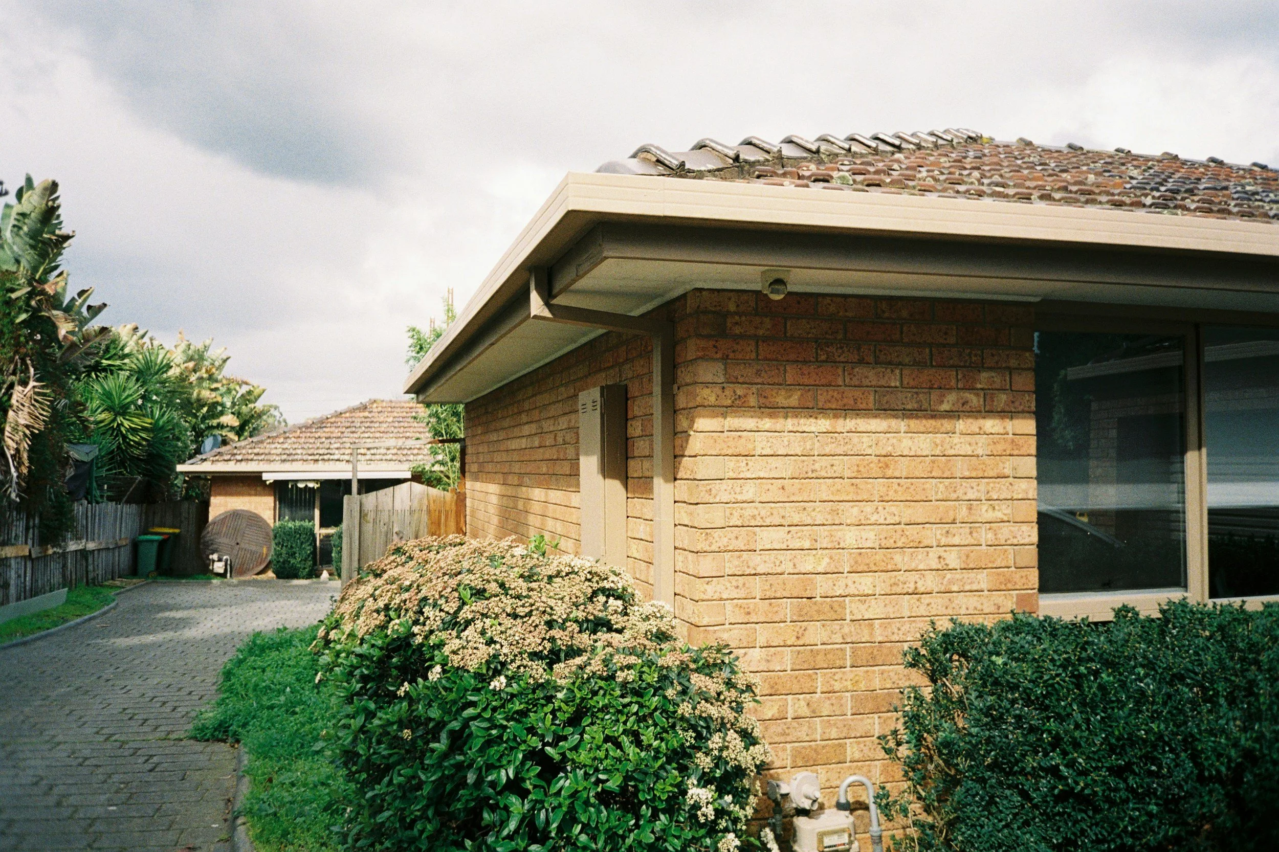 Side view of a brick house with a tiled roof, a window, garden bushes, and a paved driveway, under a cloudy sky.