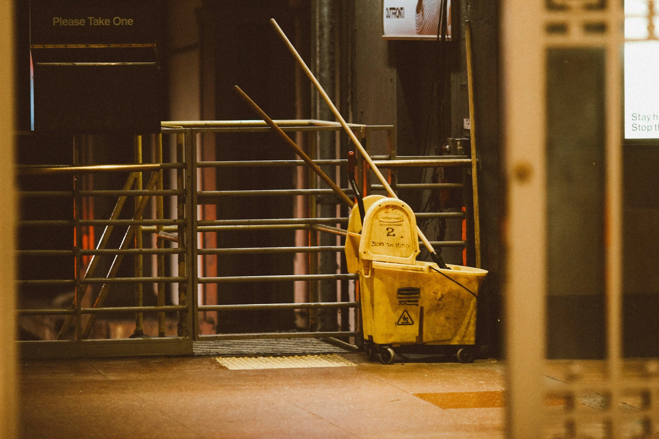 A yellow cleaning cart with mop buckets in front of a dark metal gate. A sign at the top left says "Please Take One." The background includes a concrete wall and a lit area with a sign. The floor is reddish-brown.