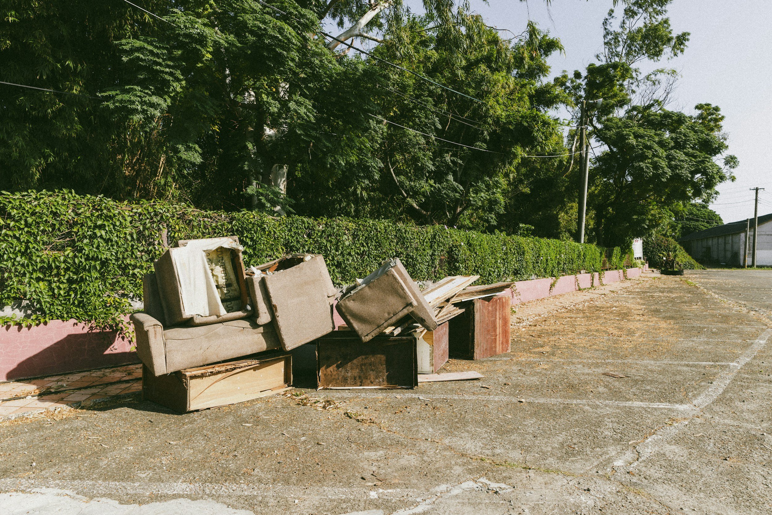 Old beige sofa and scattered furniture outdoors next to a pink planter box with green hedge. Tall trees, utility poles, and a warehouse in the background under a clear sky.