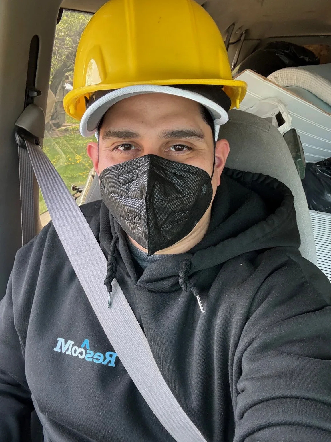 A man wearing a yellow hard hat and a black face mask taking a selfie inside a vehicle, with a grey hoodie that has blue and white text on it, and a seatbelt across his chest.