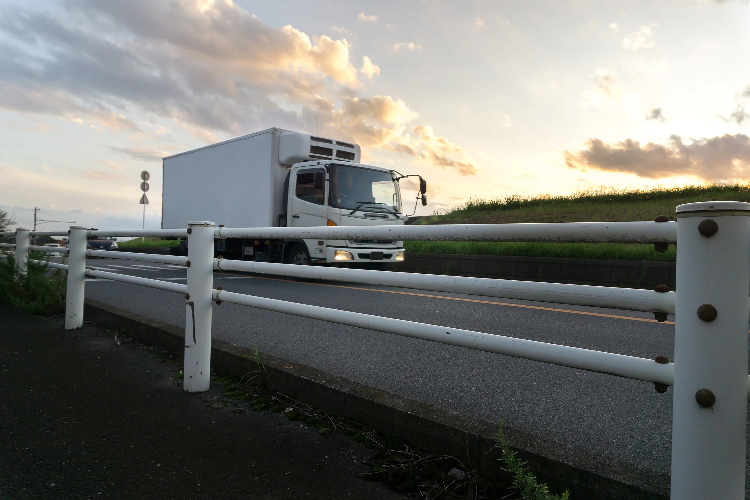 A white delivery truck driving on a highway with a guardrail in the foreground and a cloudy sunset sky in the background.