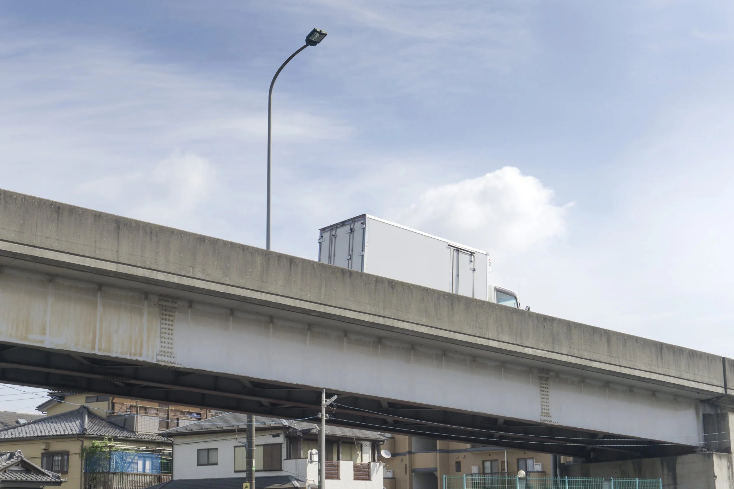 View of an elevated highway with houses and residential buildings underneath, and a streetlight in the background on a partly cloudy day.