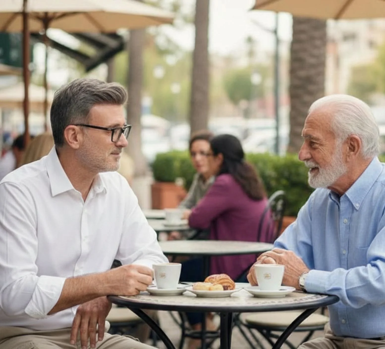 Two men having a conversation at a cafe seated across from each other with coffee cups and pastries on the table, outdoors with umbrellas and trees in the background.