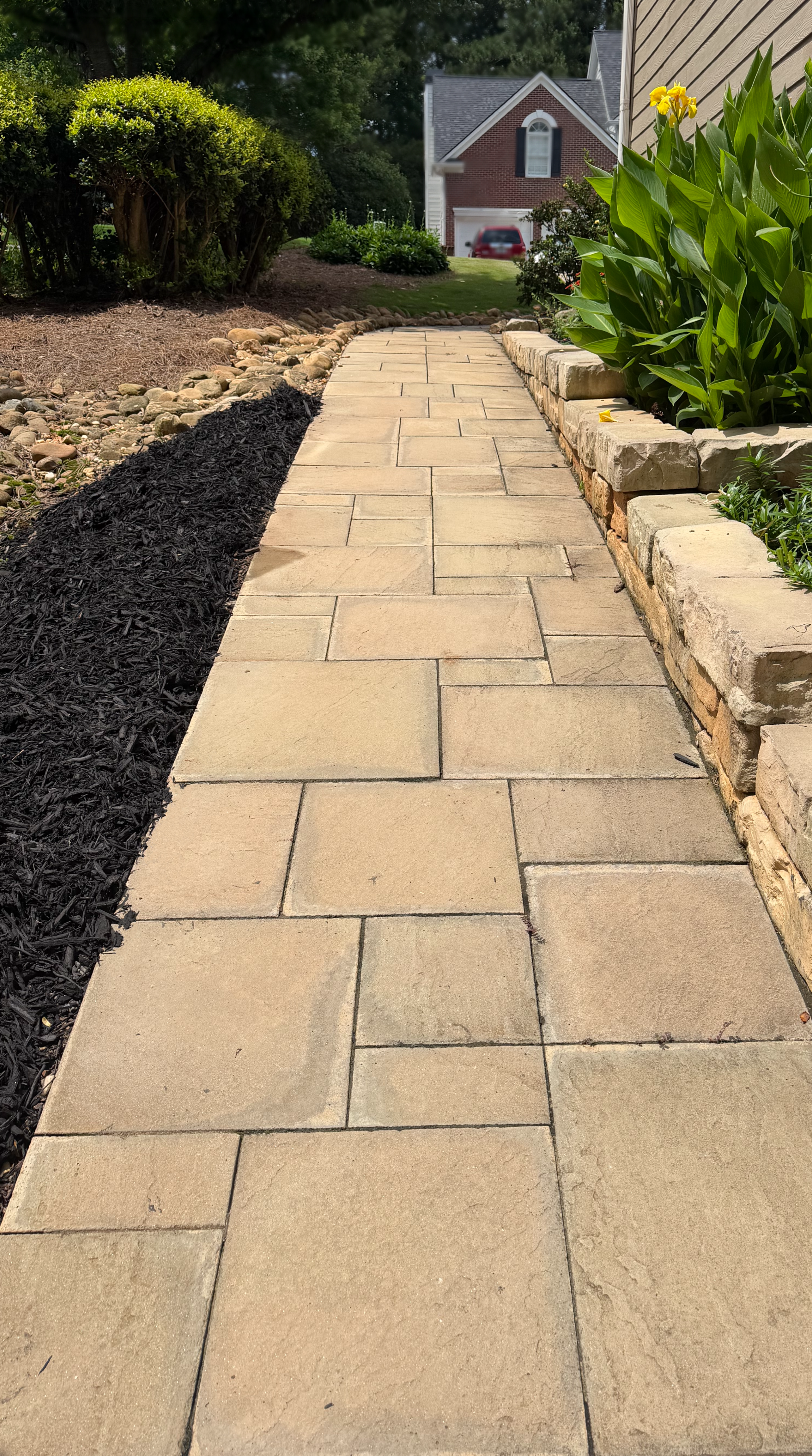 A paved walkway with beige stone pavers leading to a house with a brick facade and a garage. On the right, there is a garden bed with green leafy plants, bordered by stone blocks. On the left, there is freshly mulched soil and a shrub. In the background, there is a lawn with a car parked in the driveway and a house with black shutters.