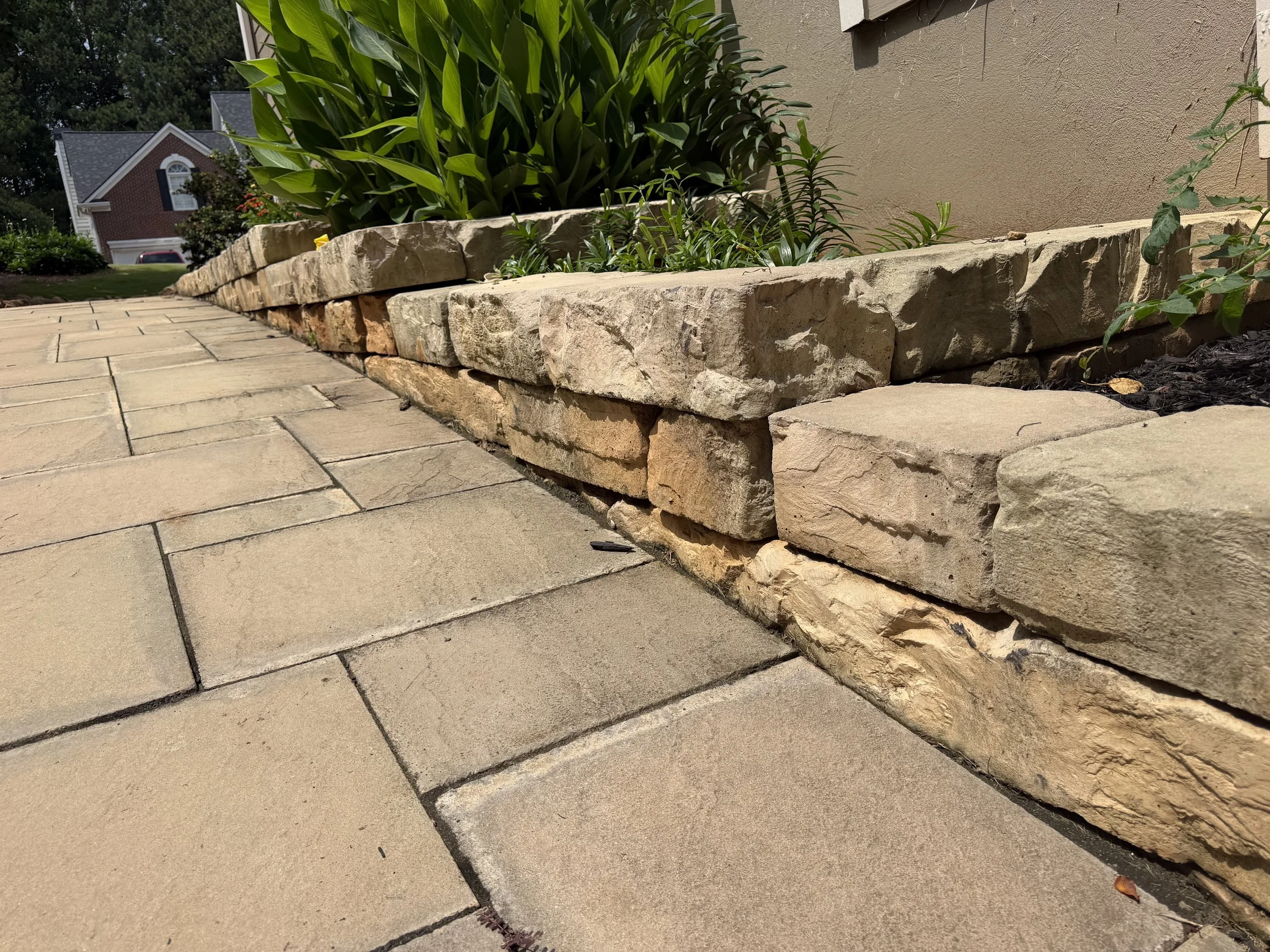 Close-up of a stone retaining wall or planter box next to a paved walkway with greenery in the background.