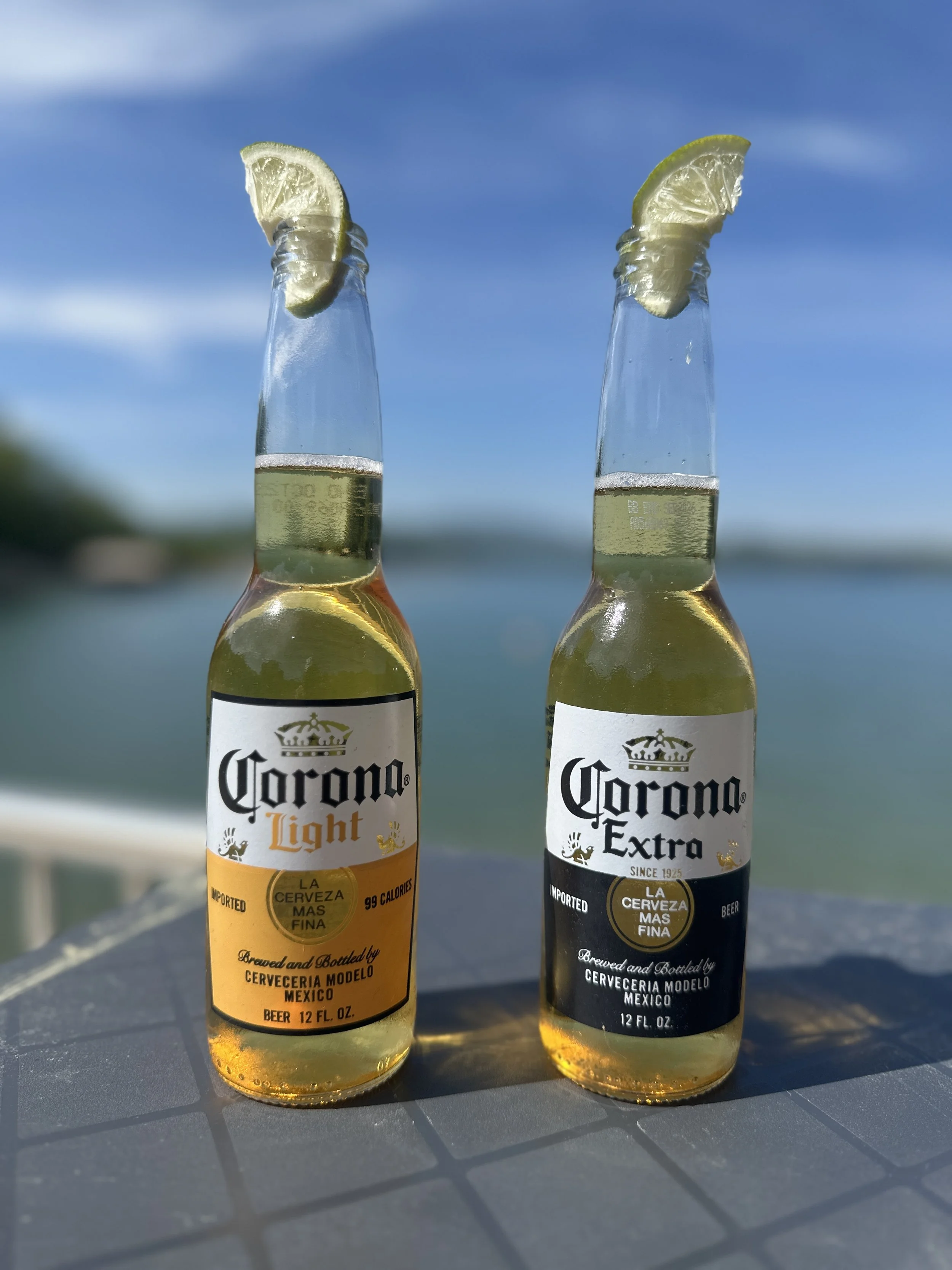 Two 12-fluid-ounce bottles of Corona beer, one Corona Light and the other Corona Extra, with lemon wedges on the necks, set against a blue sky and ocean background.