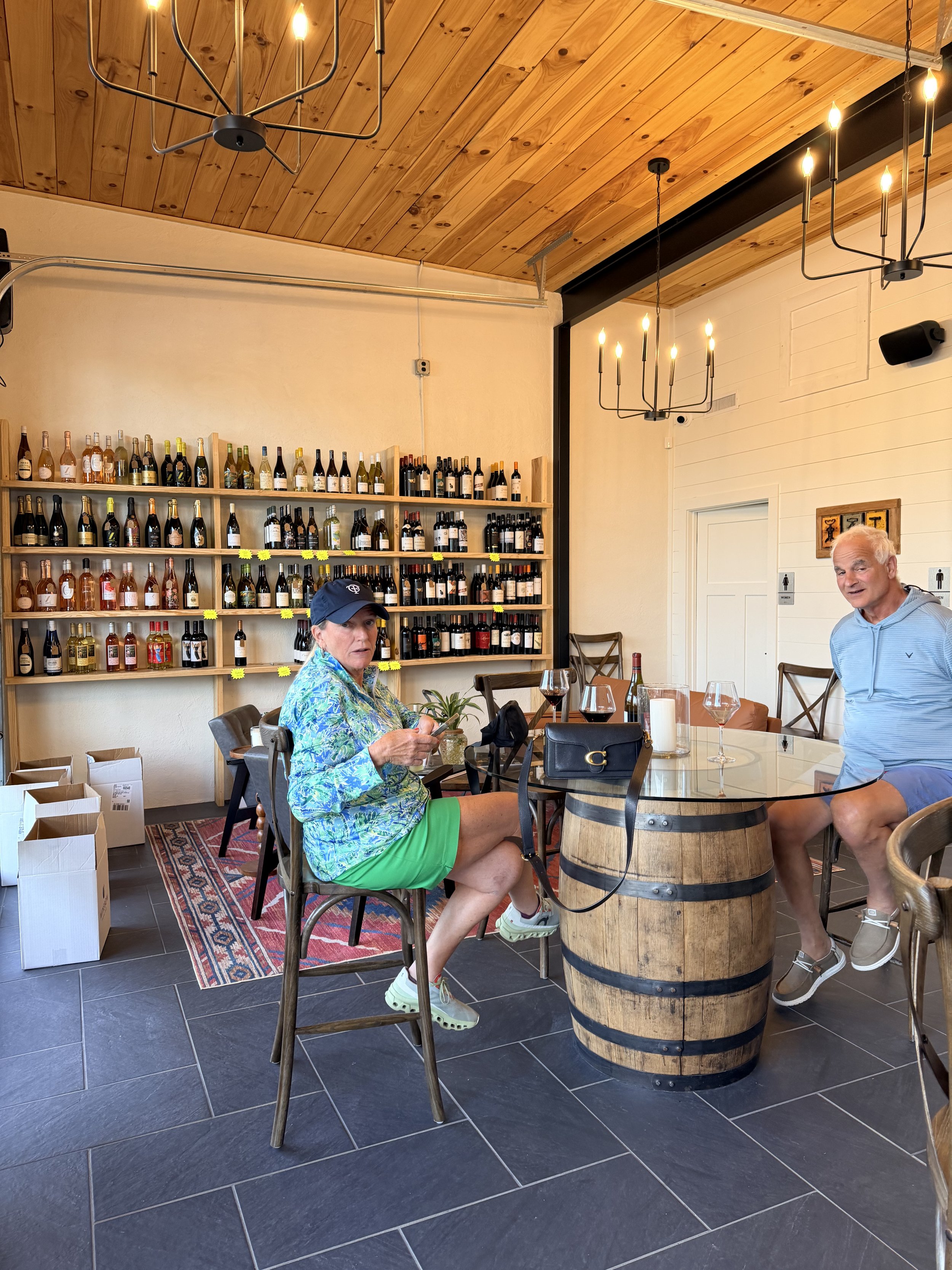 Interior of a wine shop or tasting room with wooden ceiling, chandeliers, and shelves filled with wine bottles. Two people are sitting at a table made from a wine barrel, with glasses of red wine in front of them. One woman is sitting on a chair, wearing a blue cap, floral shirt, and green shorts, looking at her phone. The man is standing, wearing a gray hoodie and shorts, and smiling. There are signs for men's and women's restrooms on the wall.