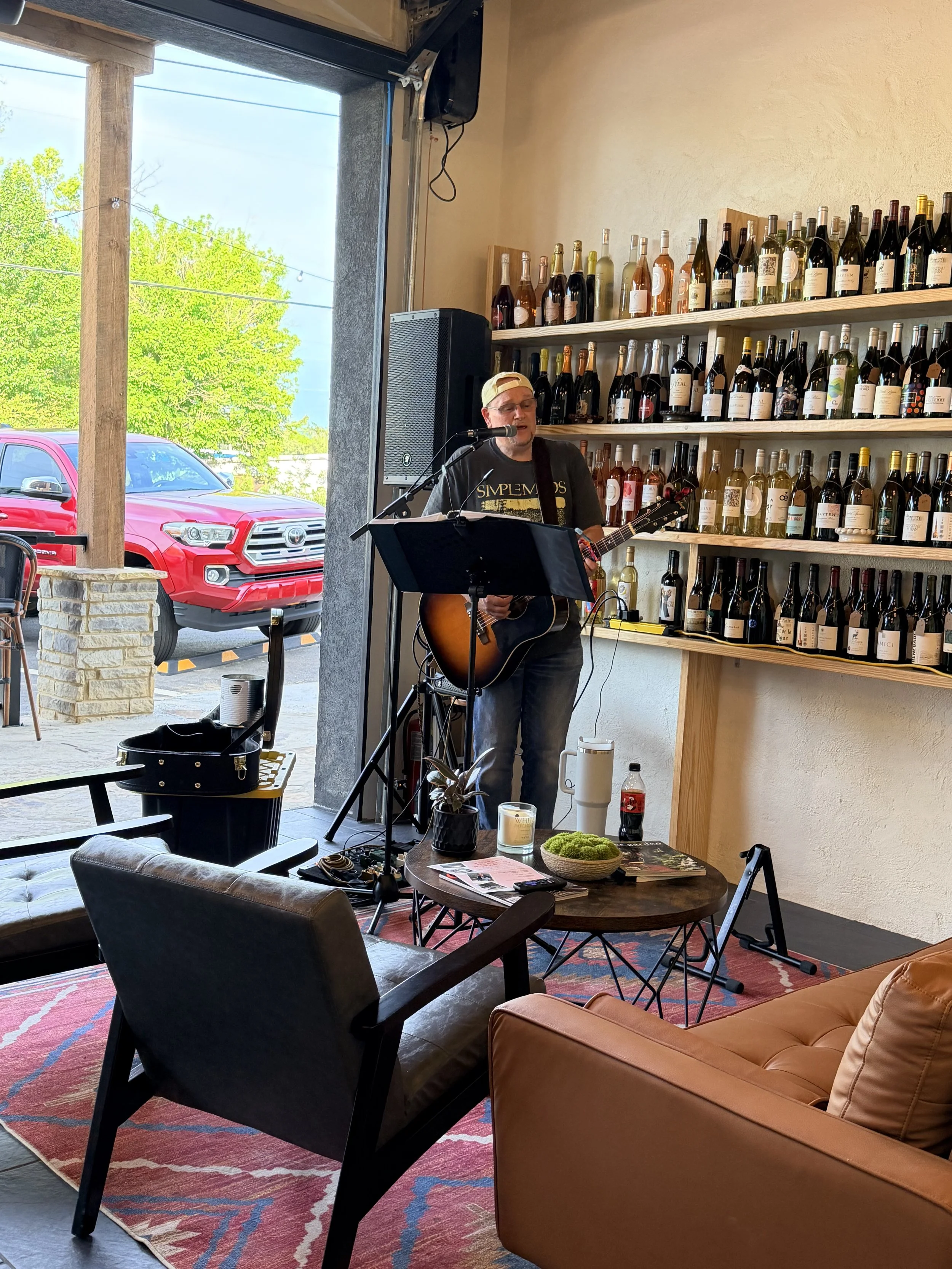 A musician wearing a casual t-shirt, jeans, and a baseball cap is playing an acoustic guitar and singing into a microphone inside a cozy cafe. The background features a large wall of wine bottles and an open garage-style door letting in natural light, with a red truck visible outside.