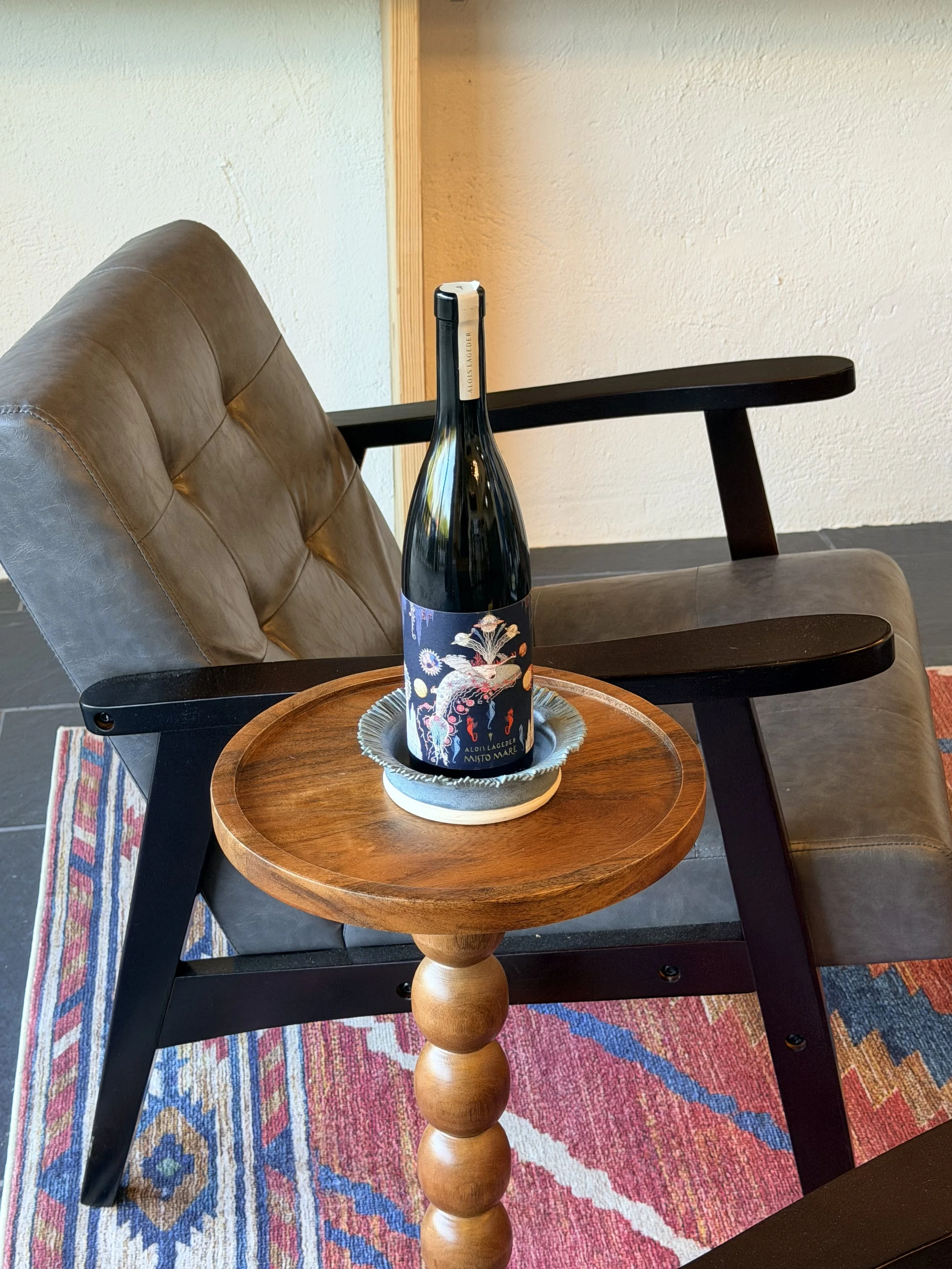 A bottle of wine with decorative label on a wooden side table next to a black chair with a leather cushion, on a colorful rug.