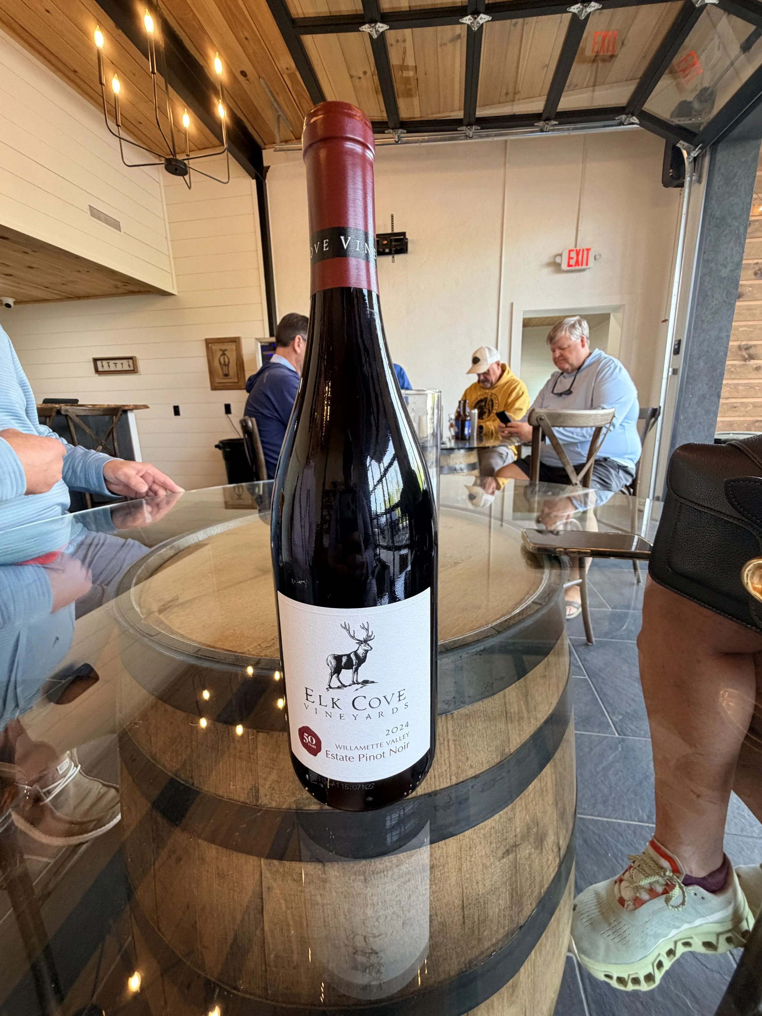 A bottle of Elk Cove Winery estate Pinot Noir wine on a glass-topped table in a restaurant, with people sitting in the background.