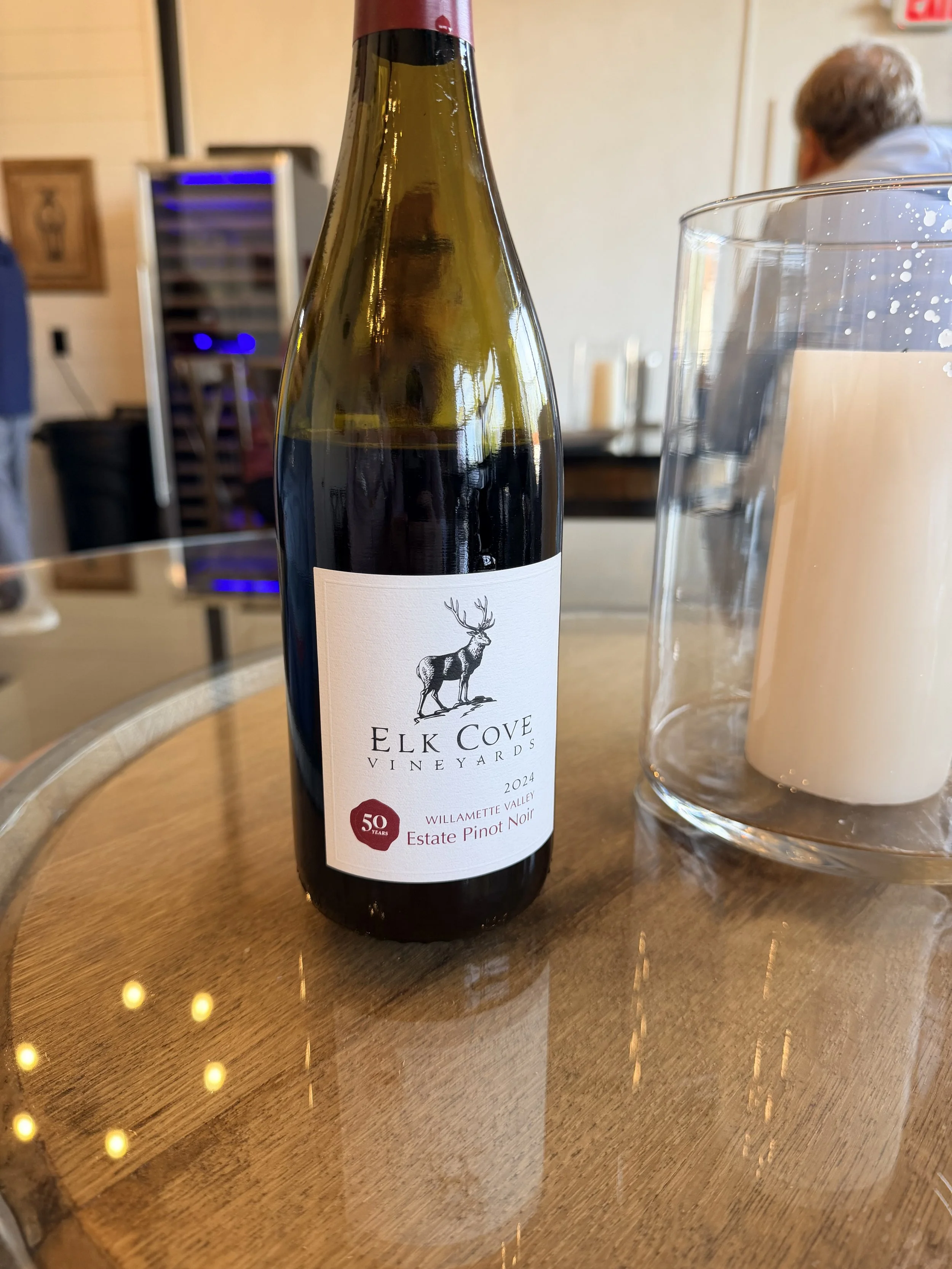 A bottle of Elk Cove Vineyards Estate Pinot Noir 2024 on a wooden table next to a glass of water, with a man and a wine cooler in the background