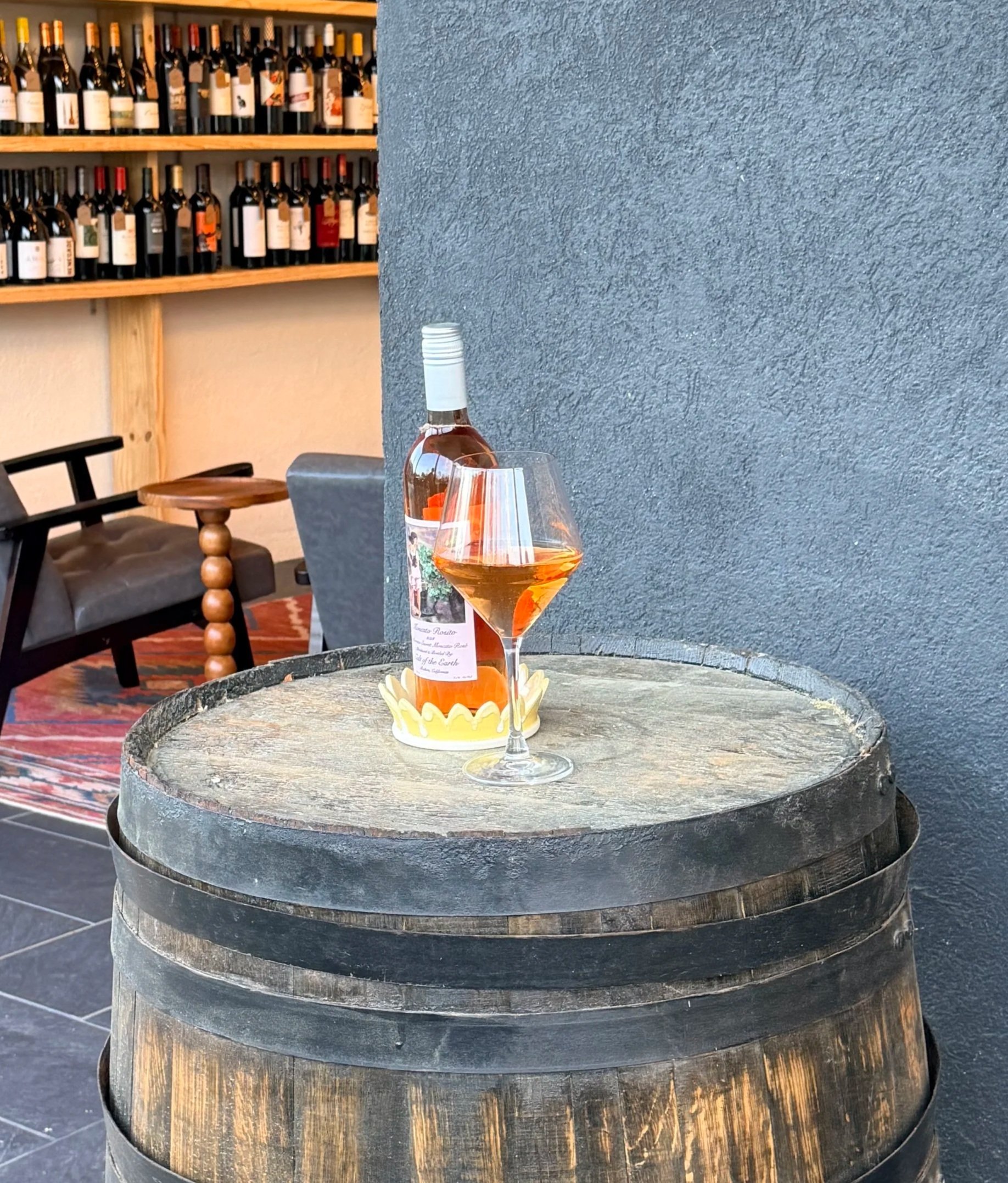 A wine bottle and a glass of rosé wine on a round wooden barrel table in a restaurant or bar setting, with shelves of wine bottles in the background.
