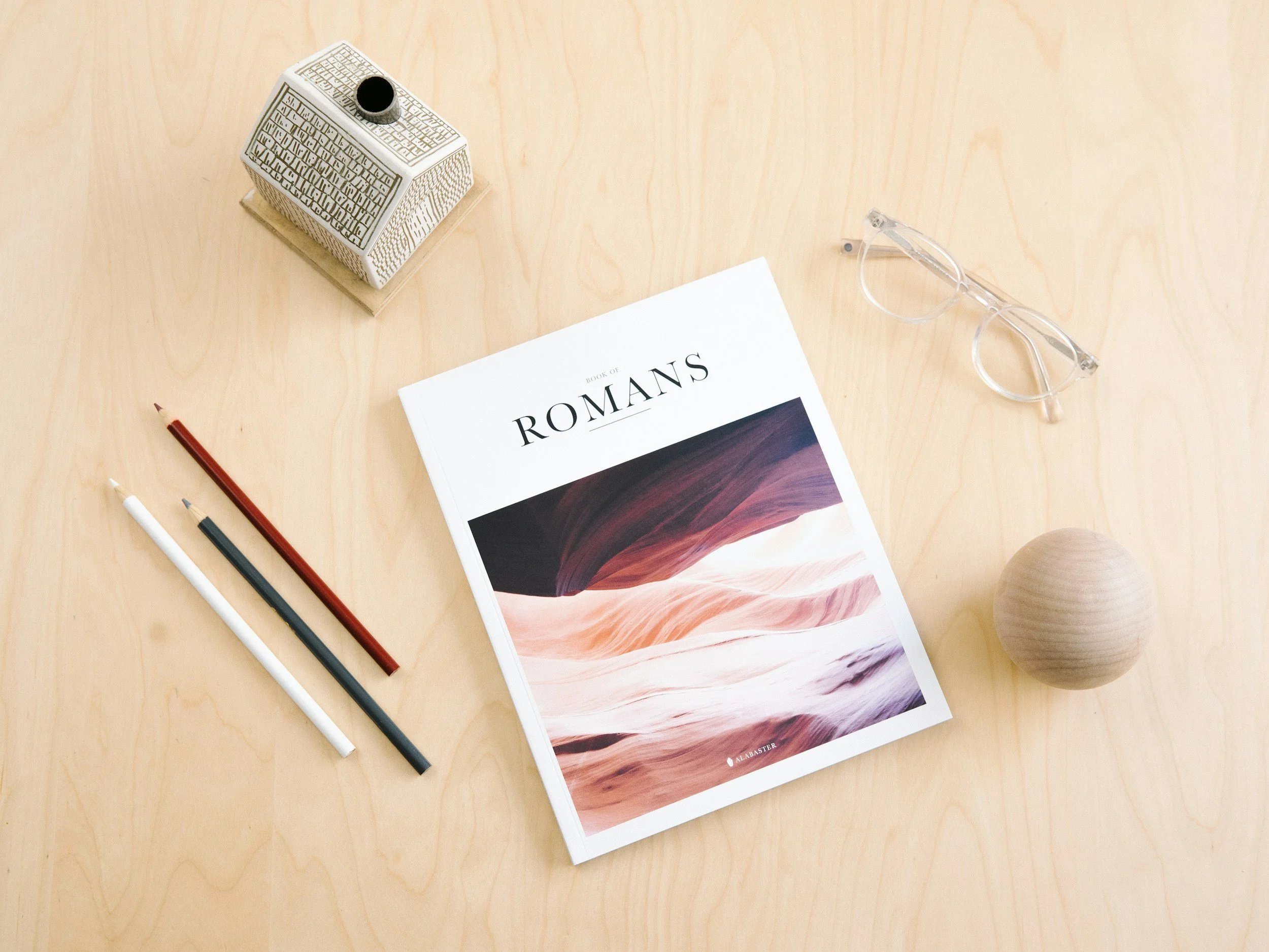 A study desk with a white book titled 'Book of Romans', eyeglasses, colored pencils, a decorative house, and a round vase on a light wood surface.