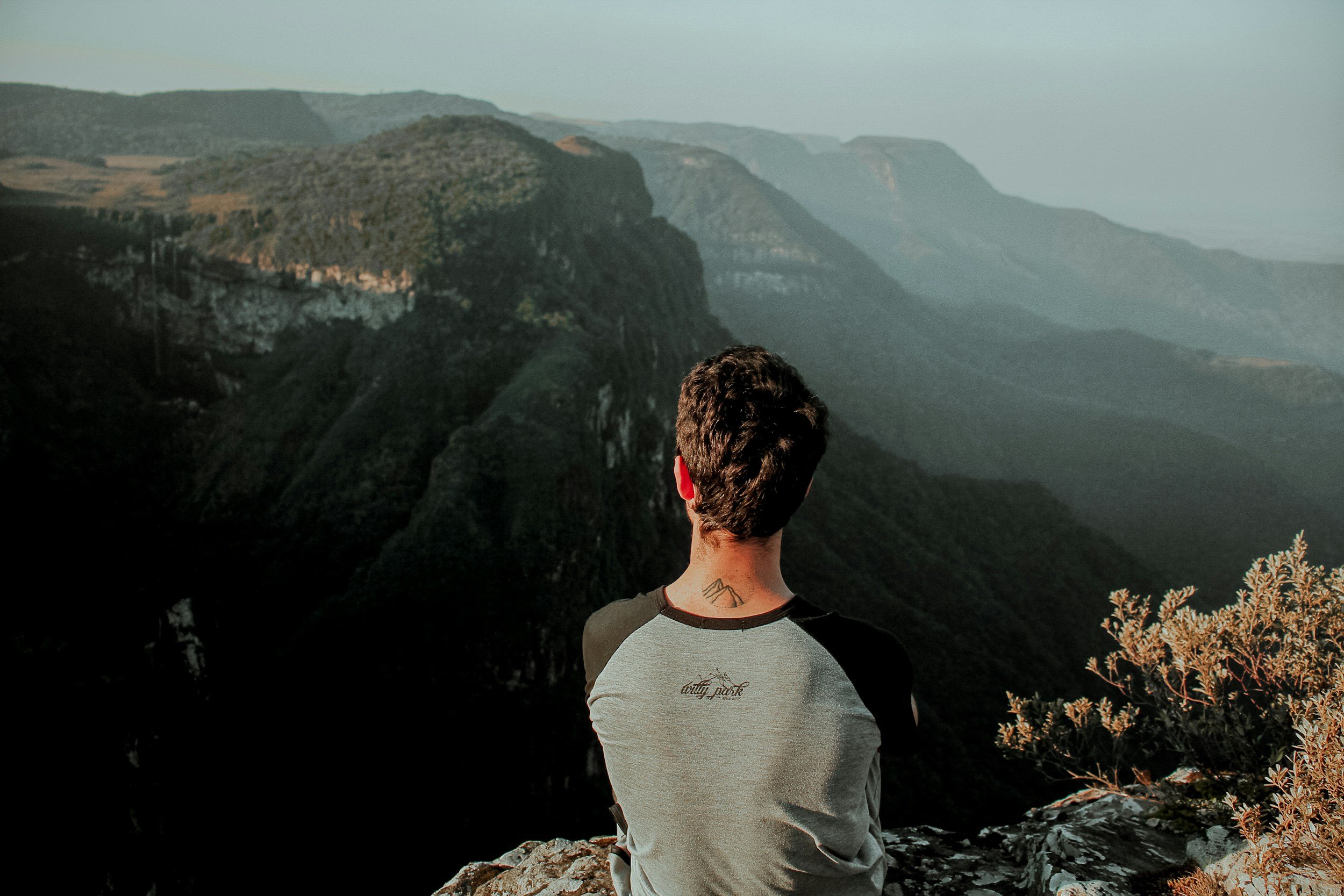 A person with short curly hair and a tattoo on the back of the neck is standing on rocky terrain, facing away from the camera, overlooking a vast landscape of mountains and valleys during daytime.