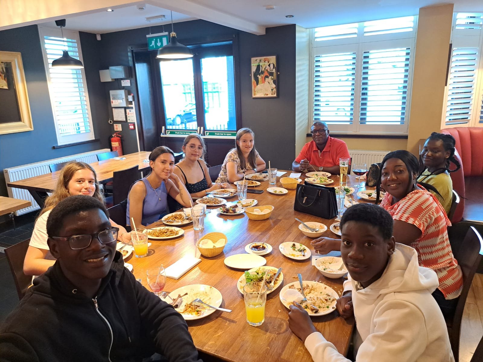 Group of people enjoying a meal together at a restaurant table with various dishes, drinks, and smiling faces.