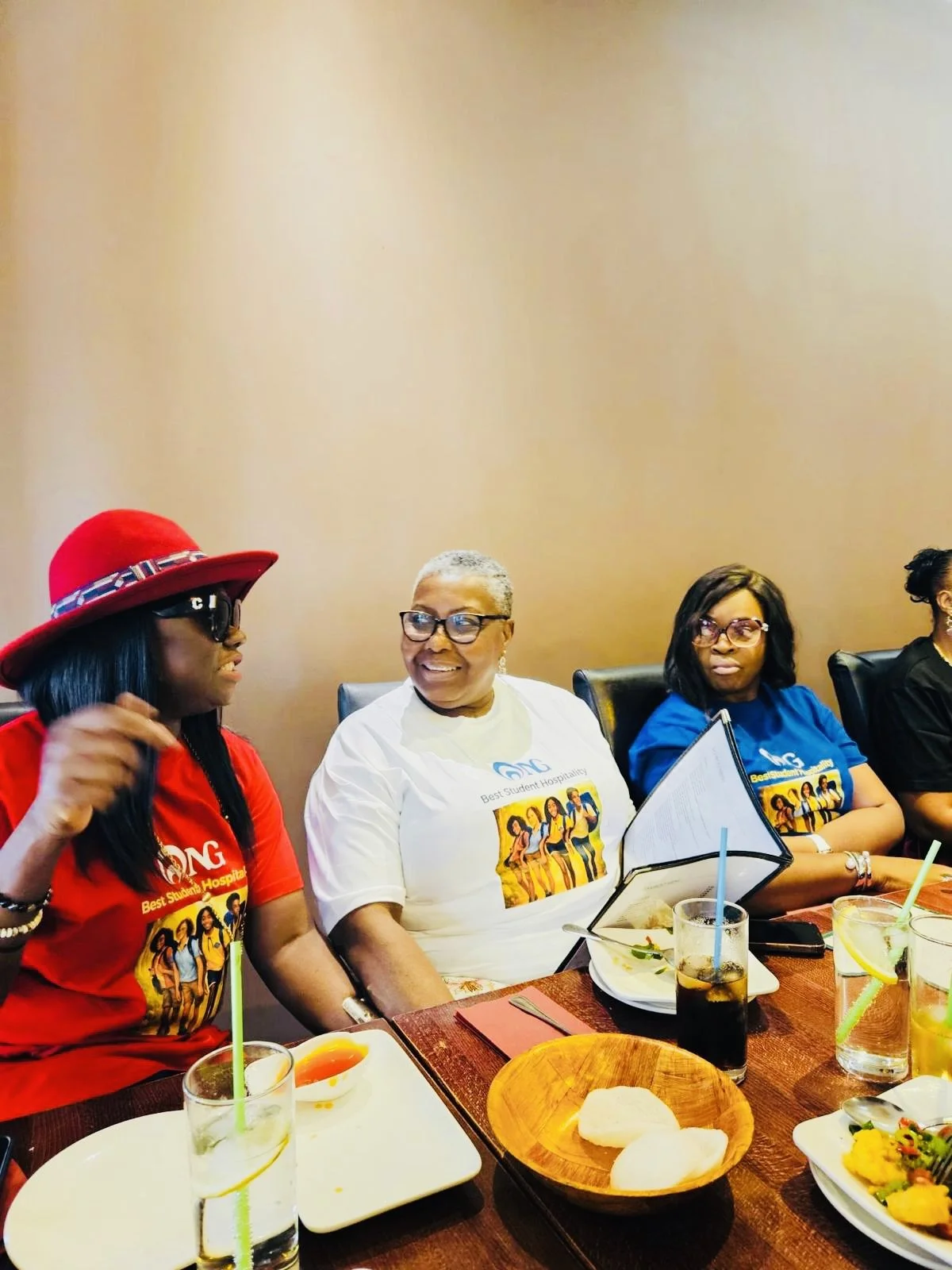 Three women sitting at a dining table during a gathering or celebration, wearing colorful T-shirts with a group photo and text, and engaging in conversation.