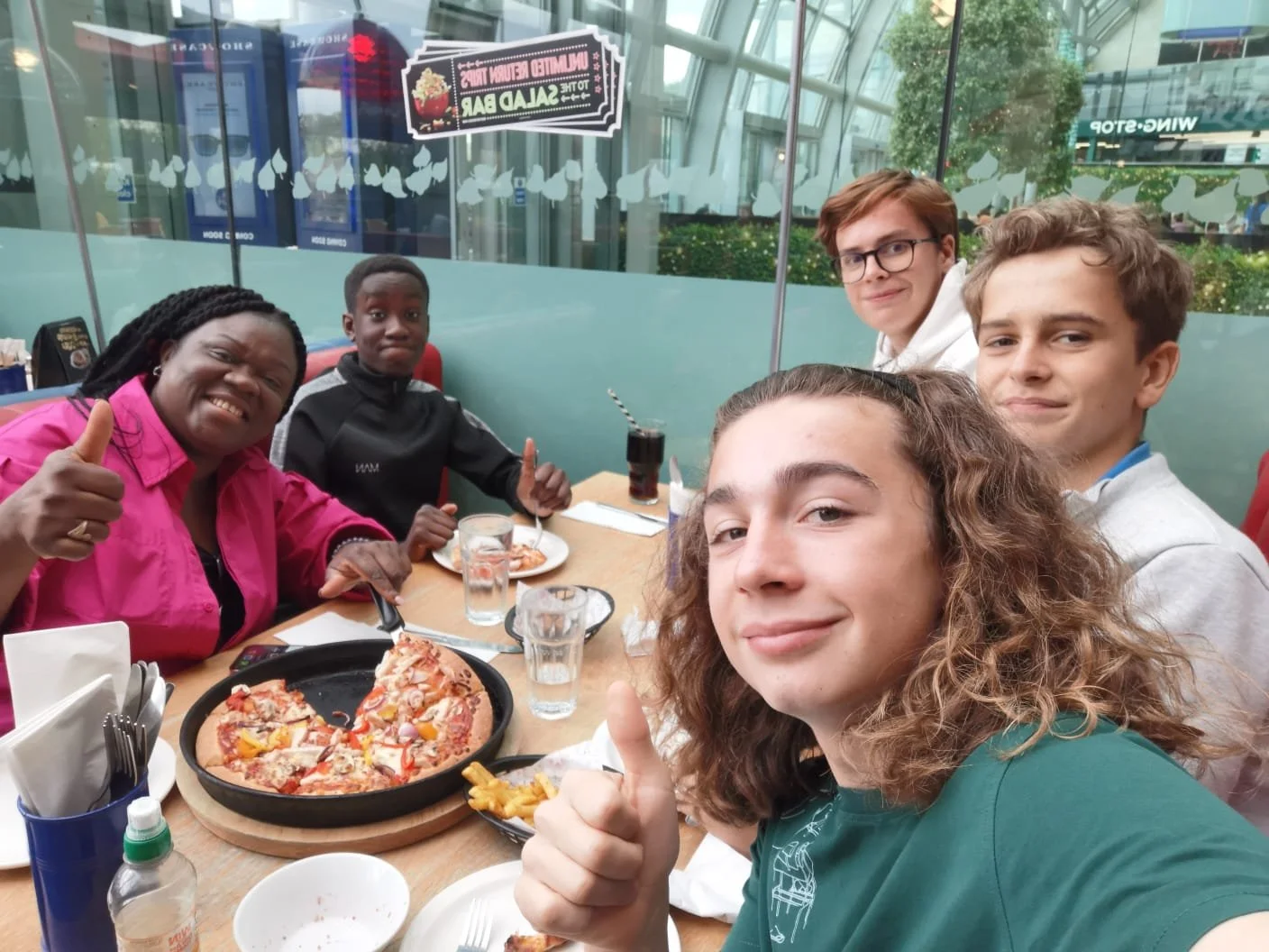 A group of five people, including two young men, two young women, and an older woman, sitting around a table in a restaurant, enjoying pizza and drinks. The woman in the foreground is giving a thumbs-up.