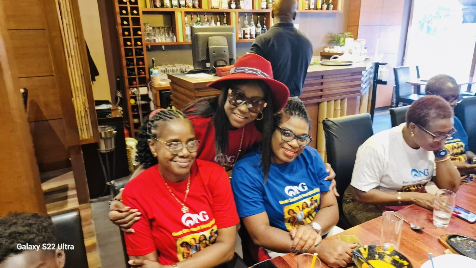Four women sitting at a dining table, smiling, with one woman standing behind them. The women are wearing colorful shirts and glasses, and there are drinks and food on the table. The background shows a bar area with bottles on shelves.